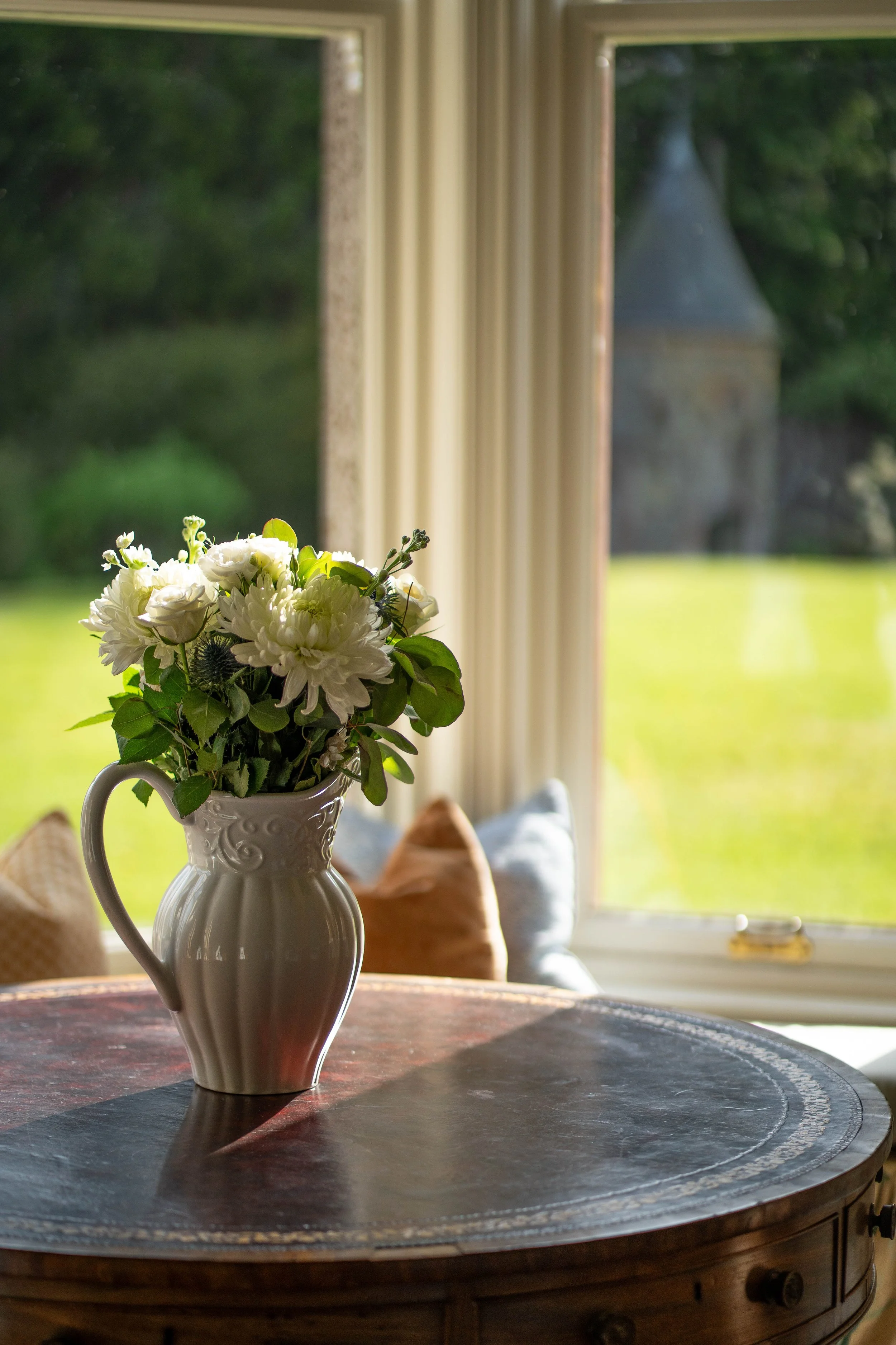 A white ceramic pitcher filled with white flowers sits on a dark wooden table in front of a window with beige curtains, with a green outdoor landscape visible through the window.
