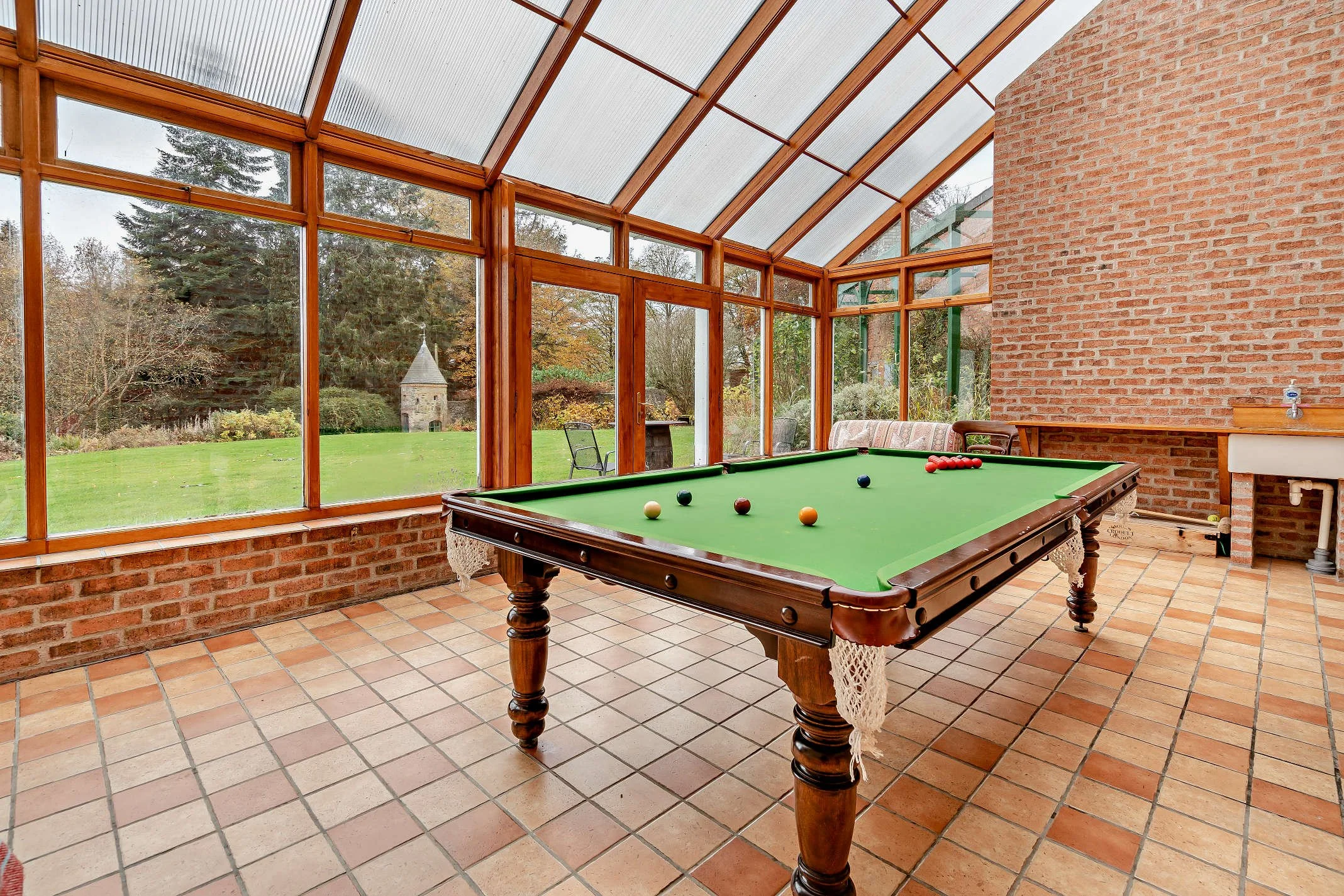 Indoor recreational room with a pool table, brick walls, large windows revealing a green lawn and trees outside, and a vaulted glass ceiling.