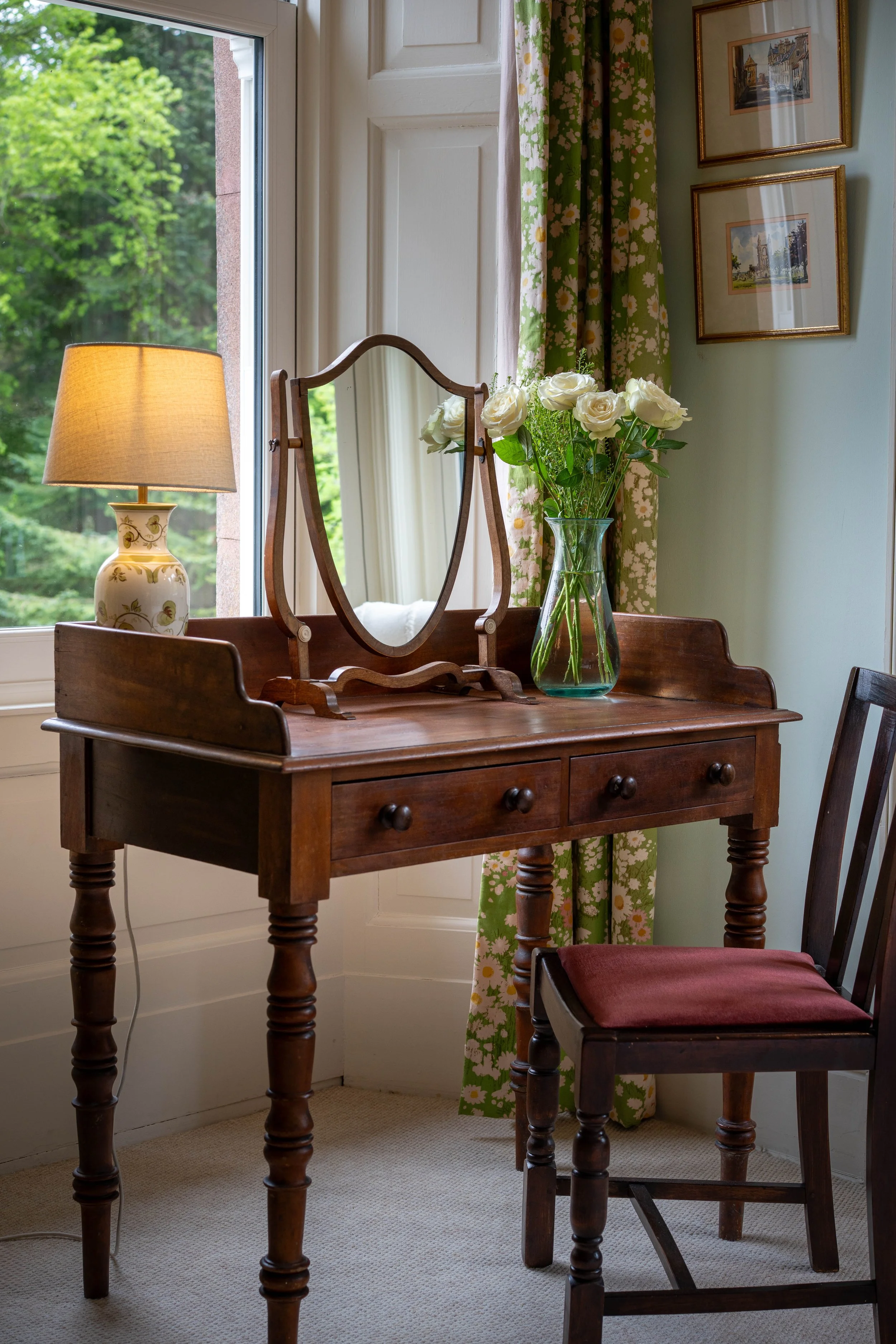 A wooden vanity table with a mirror, a vase of white roses, a table lamp, near a window with floral curtains, and a framed picture on the wall.