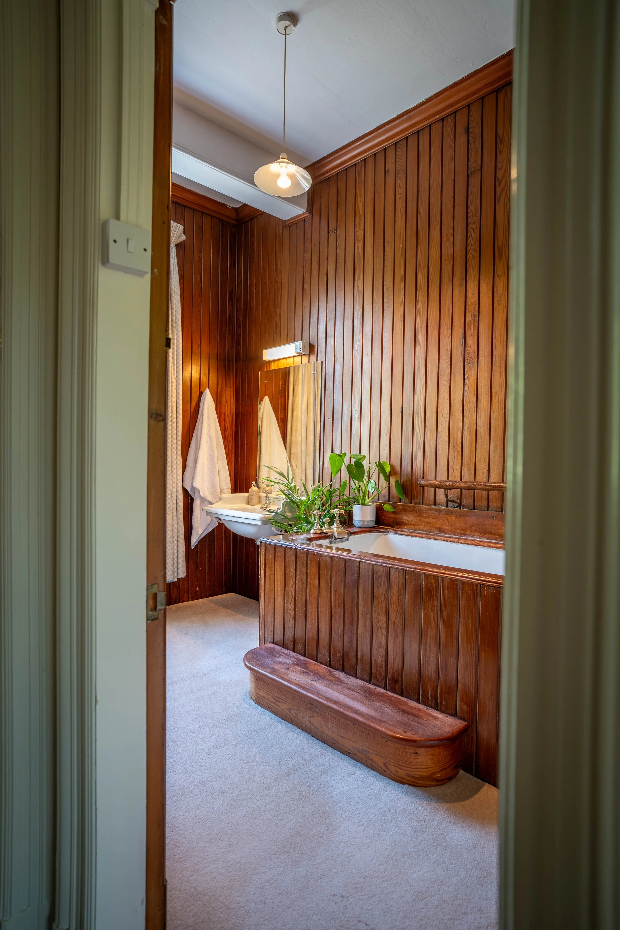 Bathroom with wood-paneled walls, a white sink with a mirror above it, a small wooden bench, and towels hanging on hooks, with a ceiling light fixture.