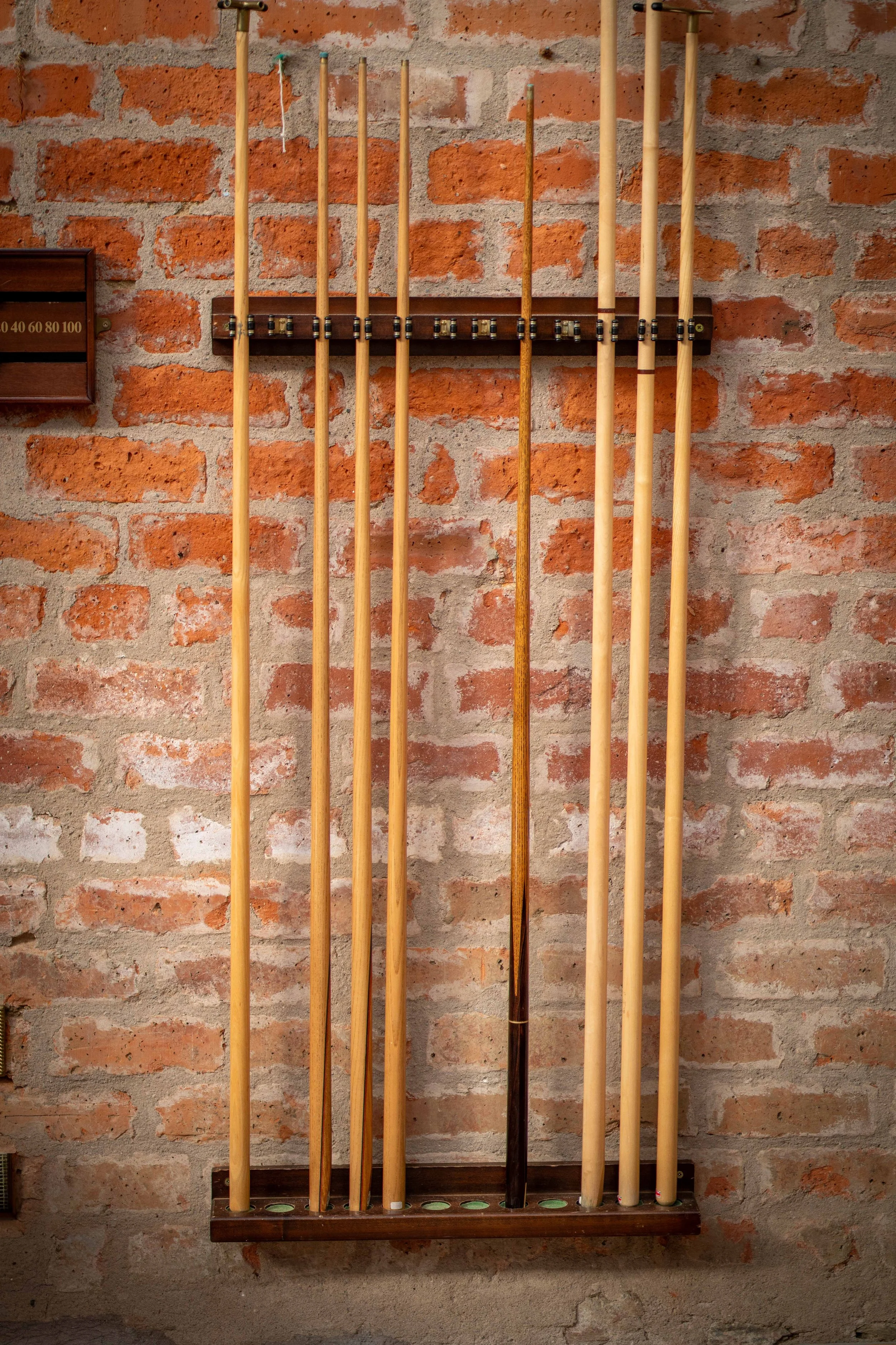 A wooden pool cue rack mounted on a brick wall, holding multiple pool cues of different lengths and colors.