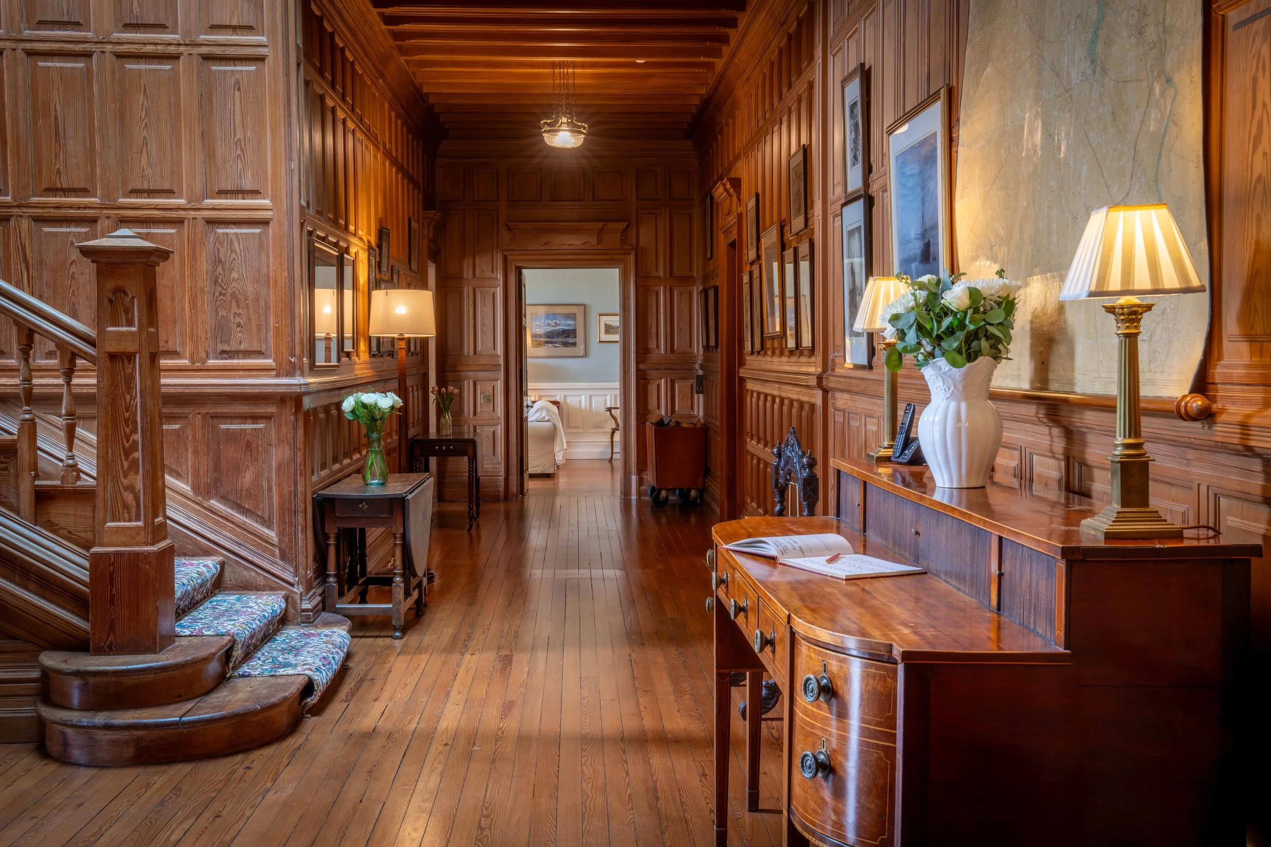 A wooden hallway with paneled walls, framed artwork, a small table with a vase of white roses, lamps, and a staircase leading to another room with lounge chairs.