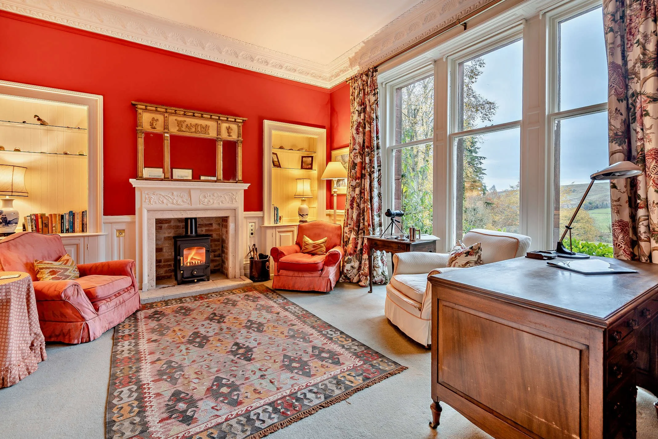 Cozy living room with red and beige walls, large windows with floral curtains, corner fireplace with a wood stove, pink velvet couch, armchair, patterned area rug, bookshelves, and a wooden desk with a lamp.