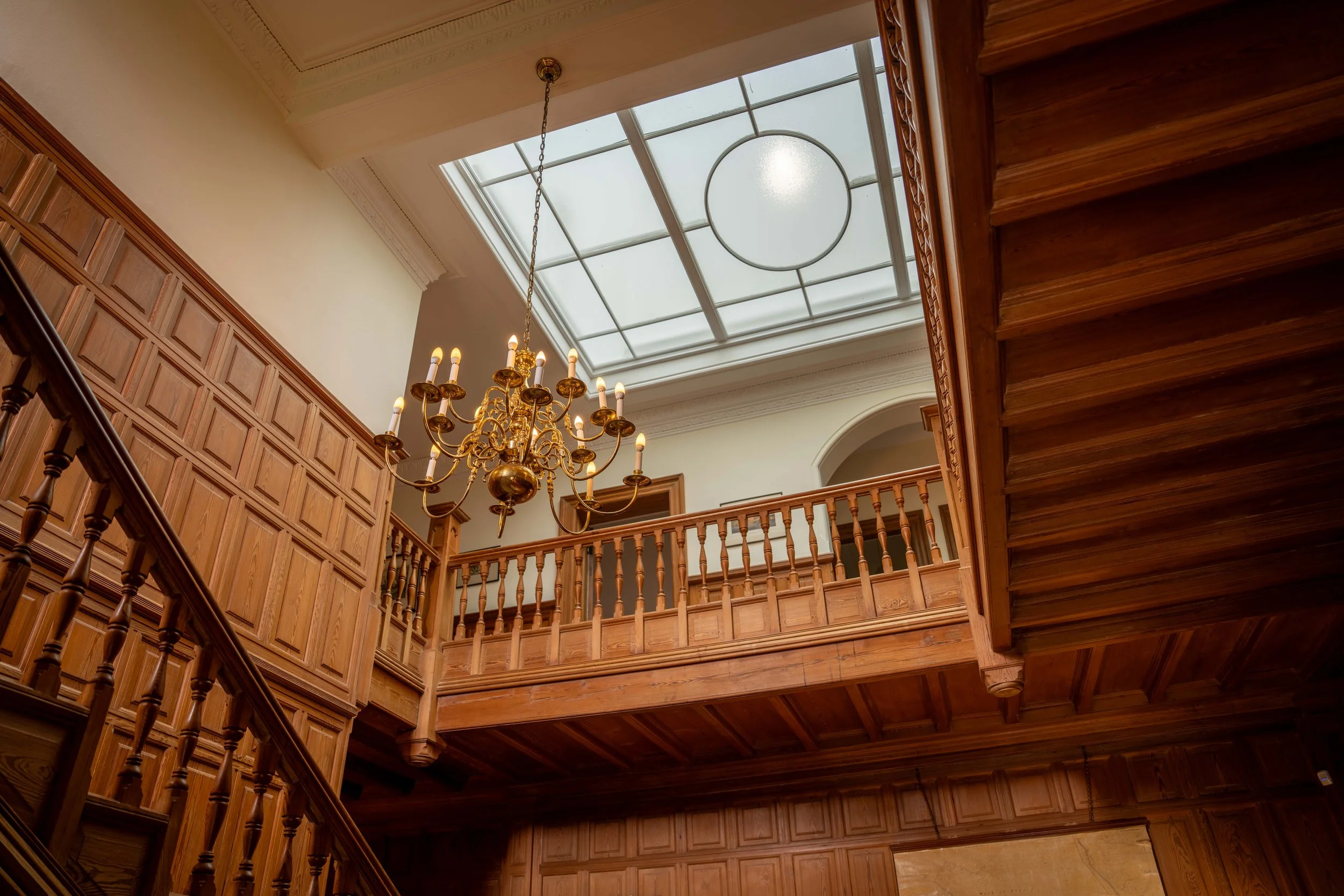 A spacious room with wooden paneling, a chandelier hanging from a ceiling with a skylight, and wooden stair railings.