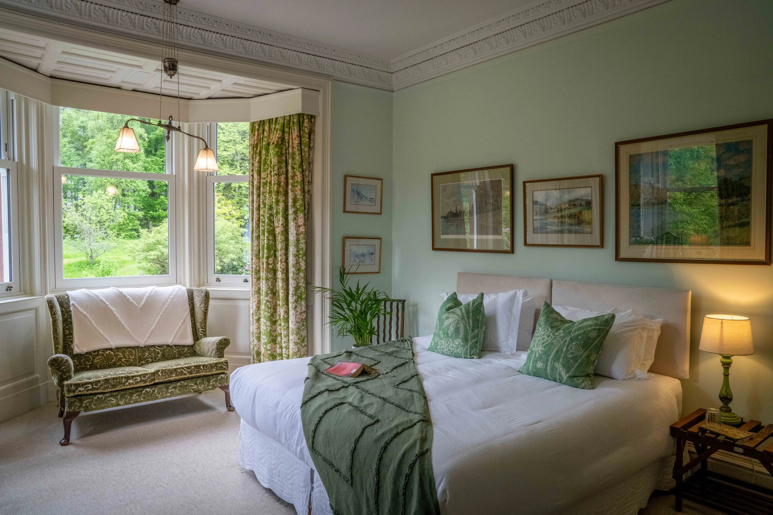 A bedroom with a large window overlooking green trees, a bed with green patterned pillows and a throw blanket, framed landscape artwork on the wall, a vintage armchair, a small side table, and leafy plants.