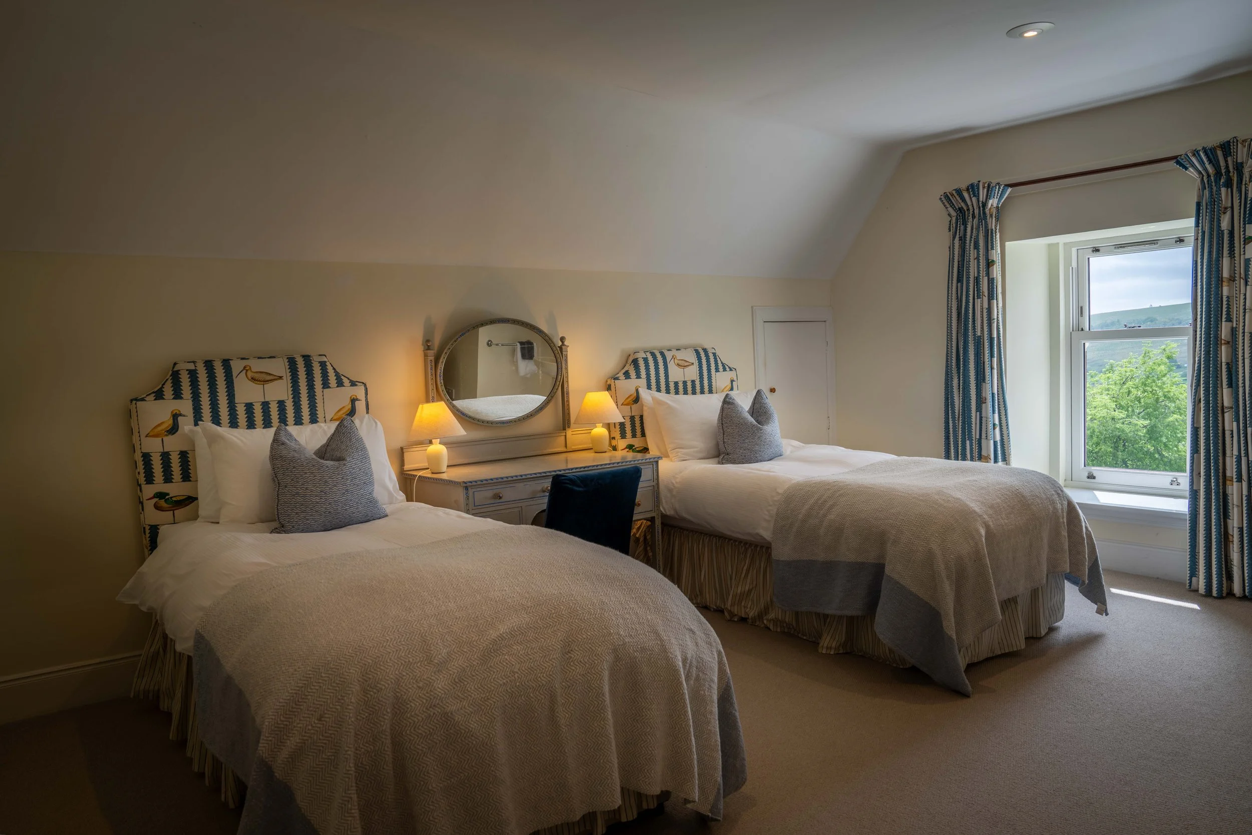 A cozy bedroom with two beds, each with tufted headboards featuring duck graphic designs, white bedding, and decorative pillows. A nightstand with two lamps and a round mirror is positioned between the beds. There is a window with blue and white patt