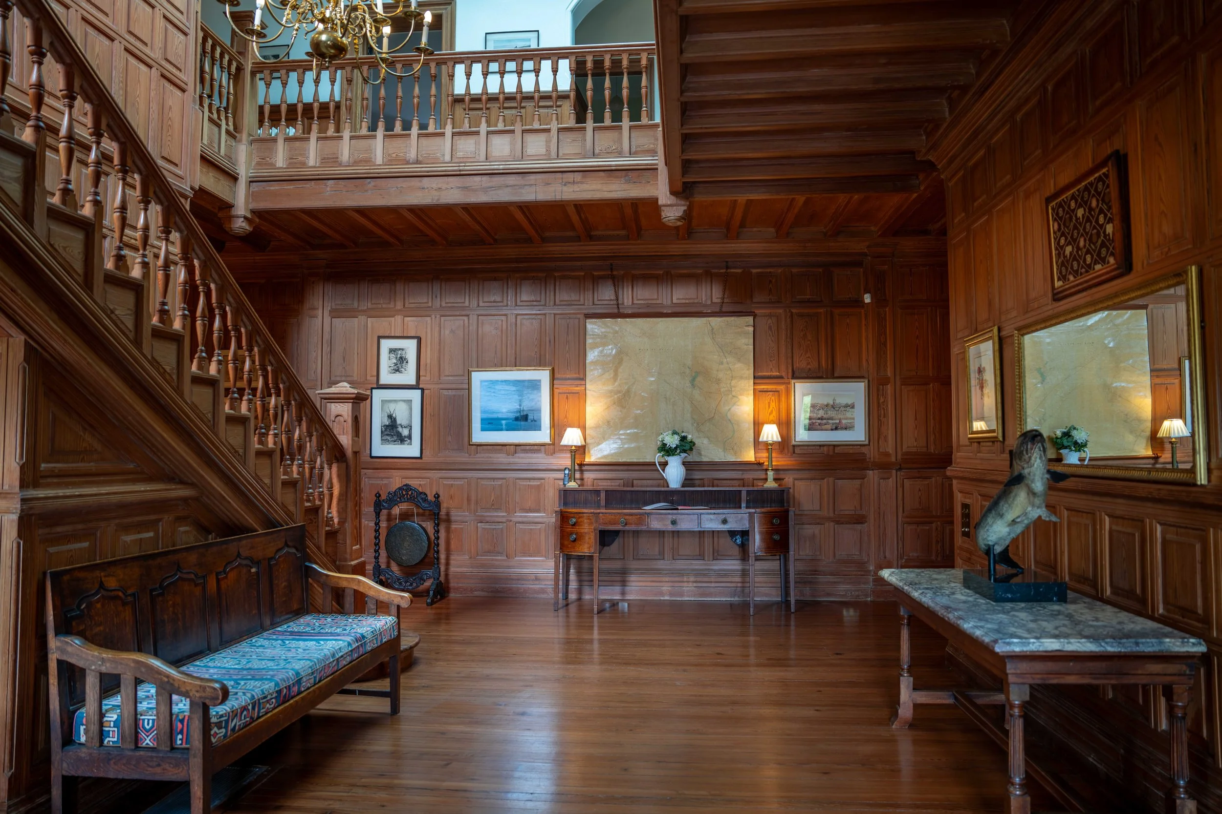 A spacious, wood-paneled room with a staircase on the left, framed artwork on the walls, a chandelier hanging from the ceiling, and brass lamps illuminating a wooden desk and decorative pieces.