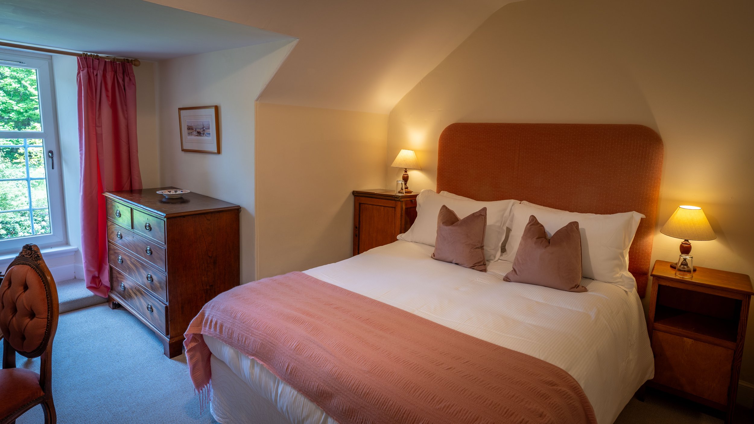 A cozy bedroom with a large bed featuring a pink bedspread and multiple pillows, bedside lamps on wooden nightstands, a wooden dresser, a window with pink curtains, and a framed picture on the wall.