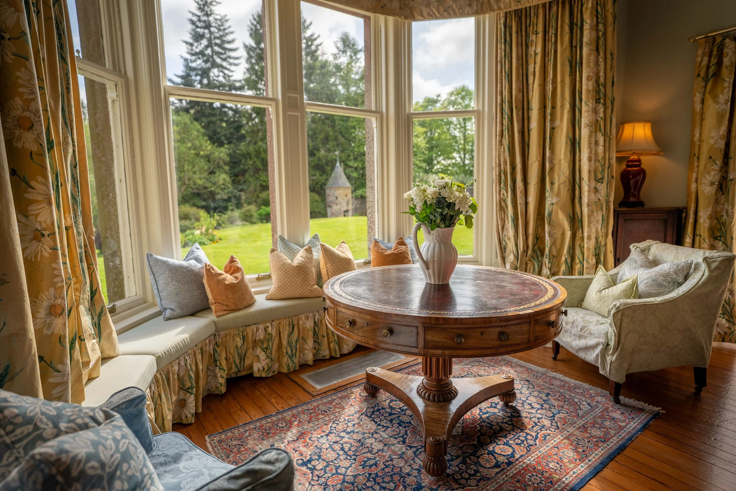A cozy living room with a large bay window and floral curtains. A round wooden table with a white vase of white flowers is in the center, surrounded by upholstered chairs and a cushioned window seat with multiple pillows. There is a rustic rug on woo