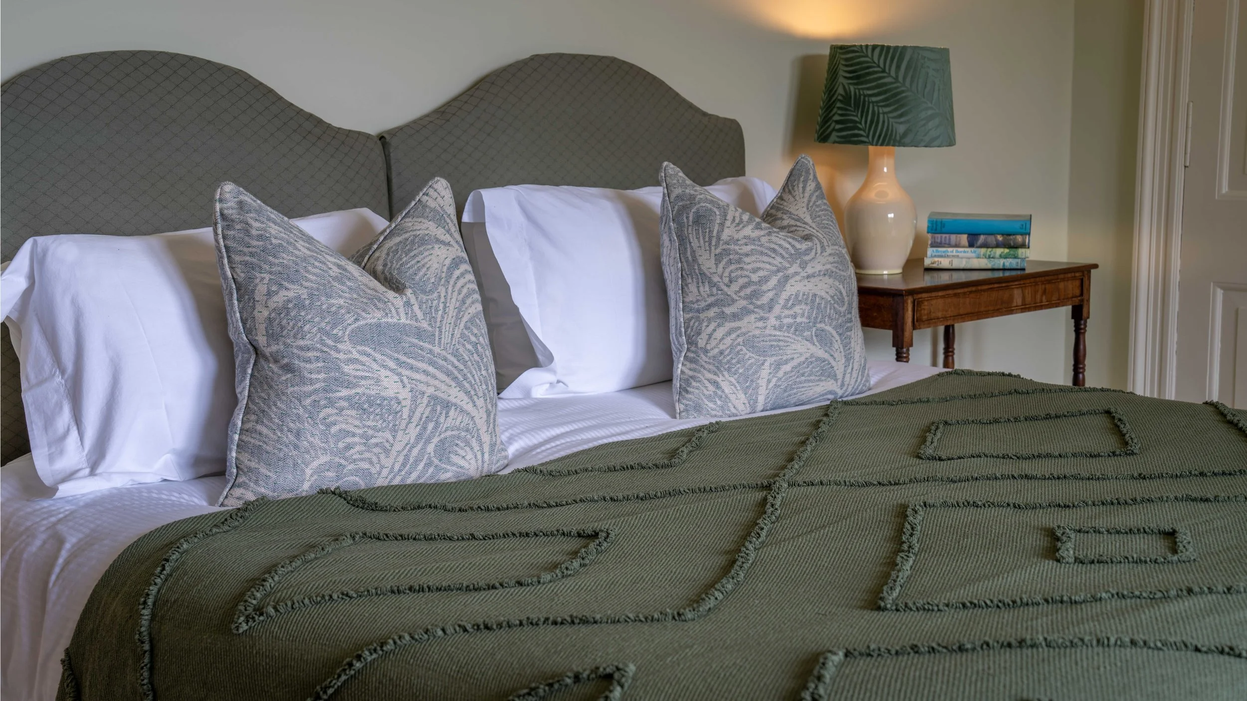 A neatly made bed with a green quilt, decorative pillows with leaf patterns, white pillowcases, and a gray upholstered headboard. Beside the bed, there is a wooden nightstand with a stack of books, a beige ceramic lamp with a green leafy lampshade, a