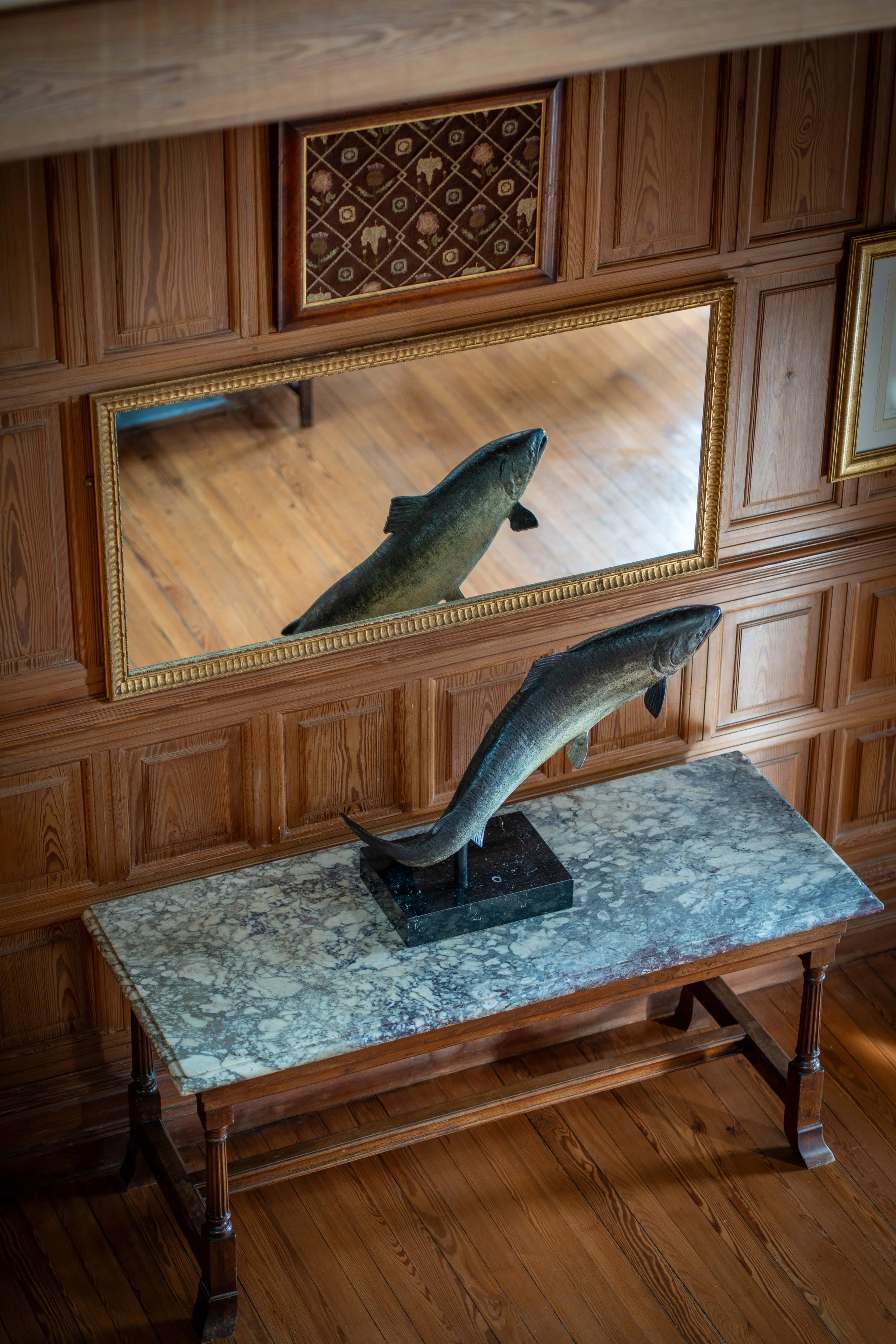 Wood-paneled wall with framed artwork, a mirror, and a taxidermy fish mounted on a stand on a marble-topped wooden table.