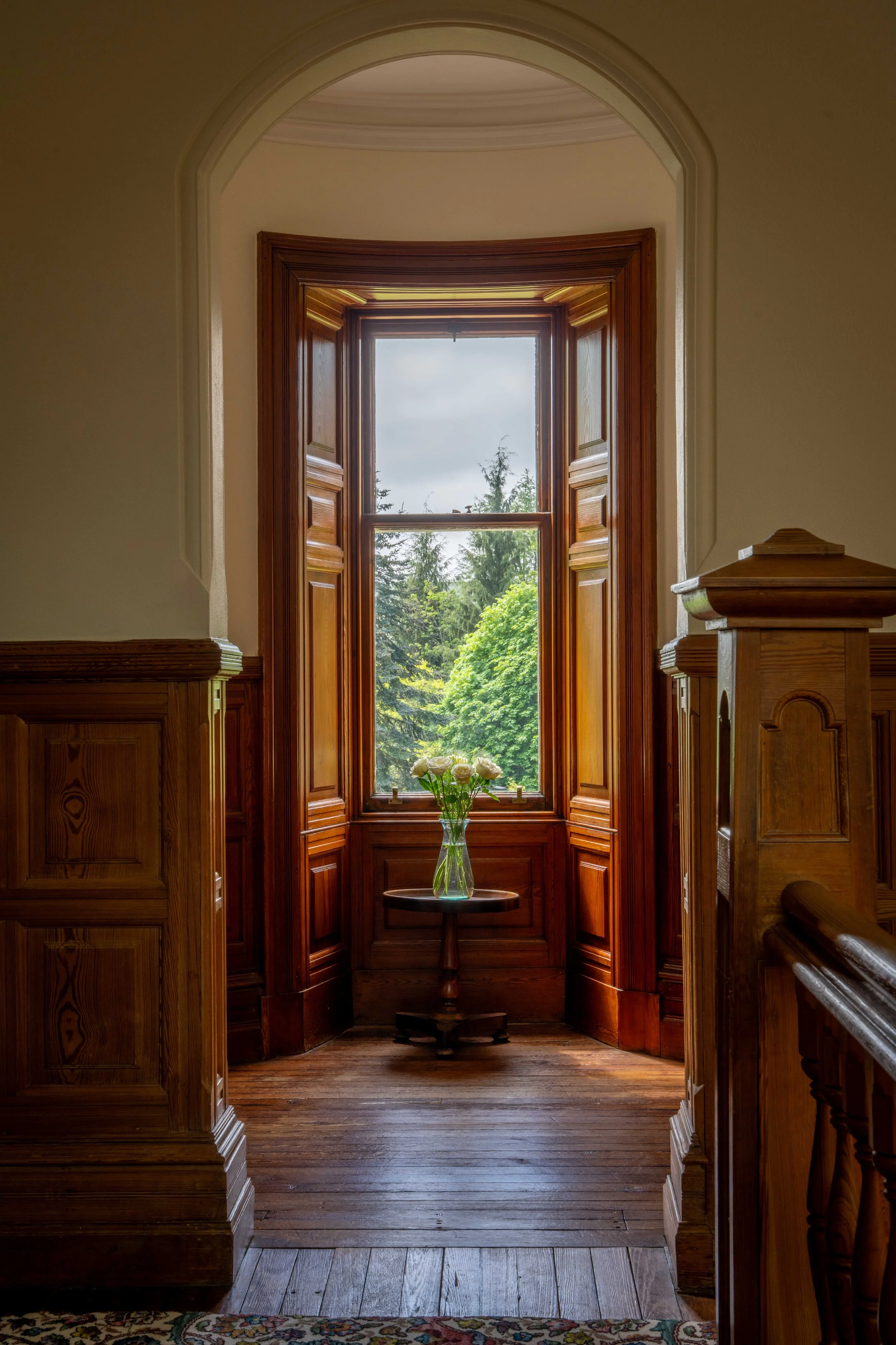 A wooden-framed window with open shutters, overlooking a lush green landscape with trees; a small round table with a glass vase of white roses is positioned in front of the window inside a room with wooden paneling.