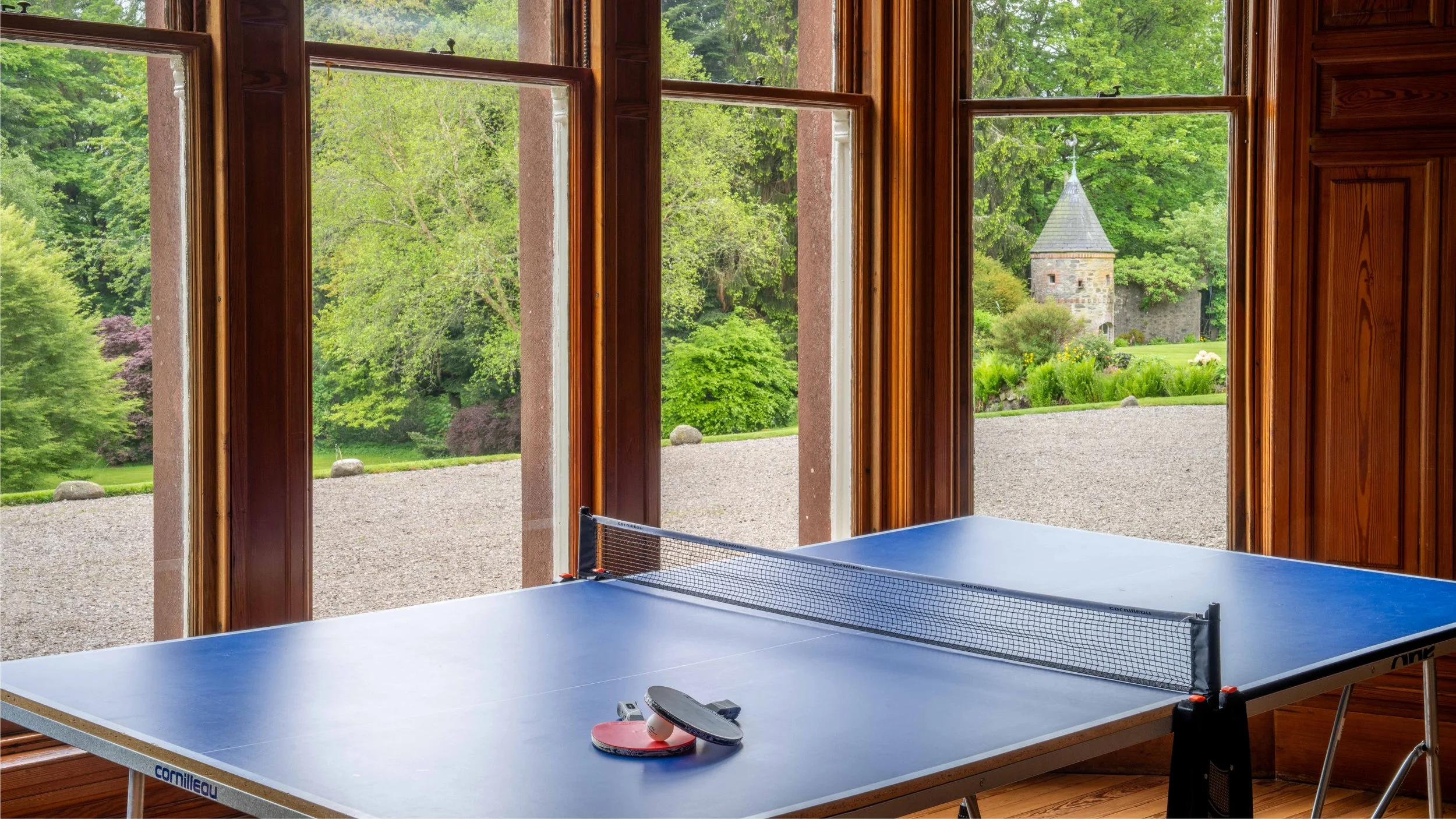 Indoor room with a table tennis table near large windows showing a lush green outdoor landscape with trees, a gravel path, and a stone tower in the distance.