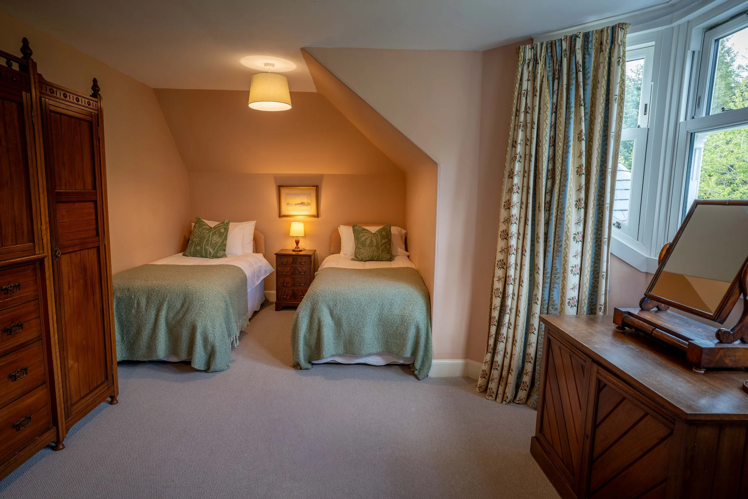 A cozy bedroom with two twin beds, green bedspreads, and matching pillows, separated by a nightstand with a lamp. There is a framed picture above the nightstand. To the left, there is a wooden wardrobe, and to the right, a window with floral curtains