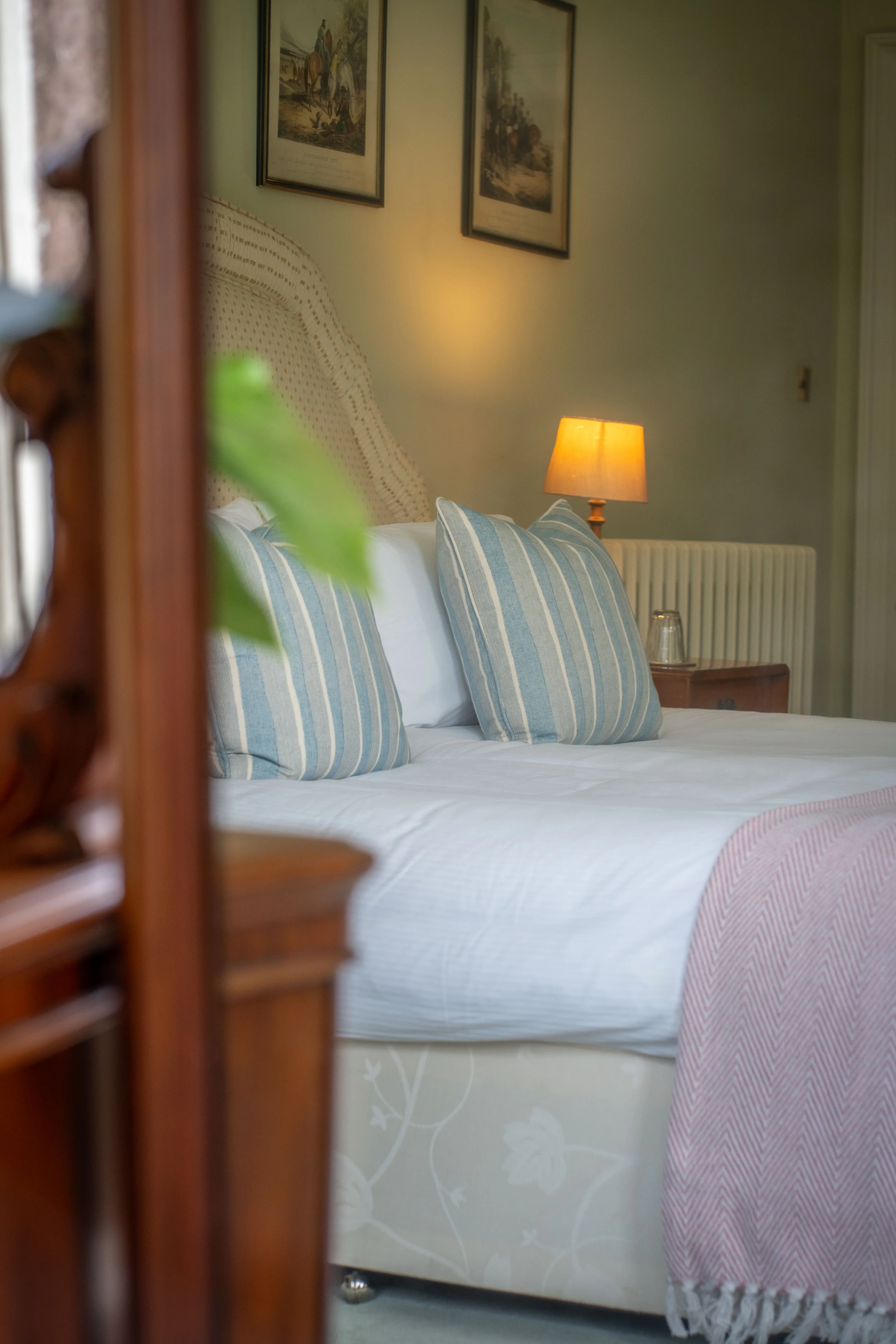 A glimpse of a bedroom through a partially open wooden louvered door. The bed has white sheets, blue and white striped pillows, a beige headboard, and a pink and white blanket. There is a bedside table with a yellow lampshade, glass, and a radiator o