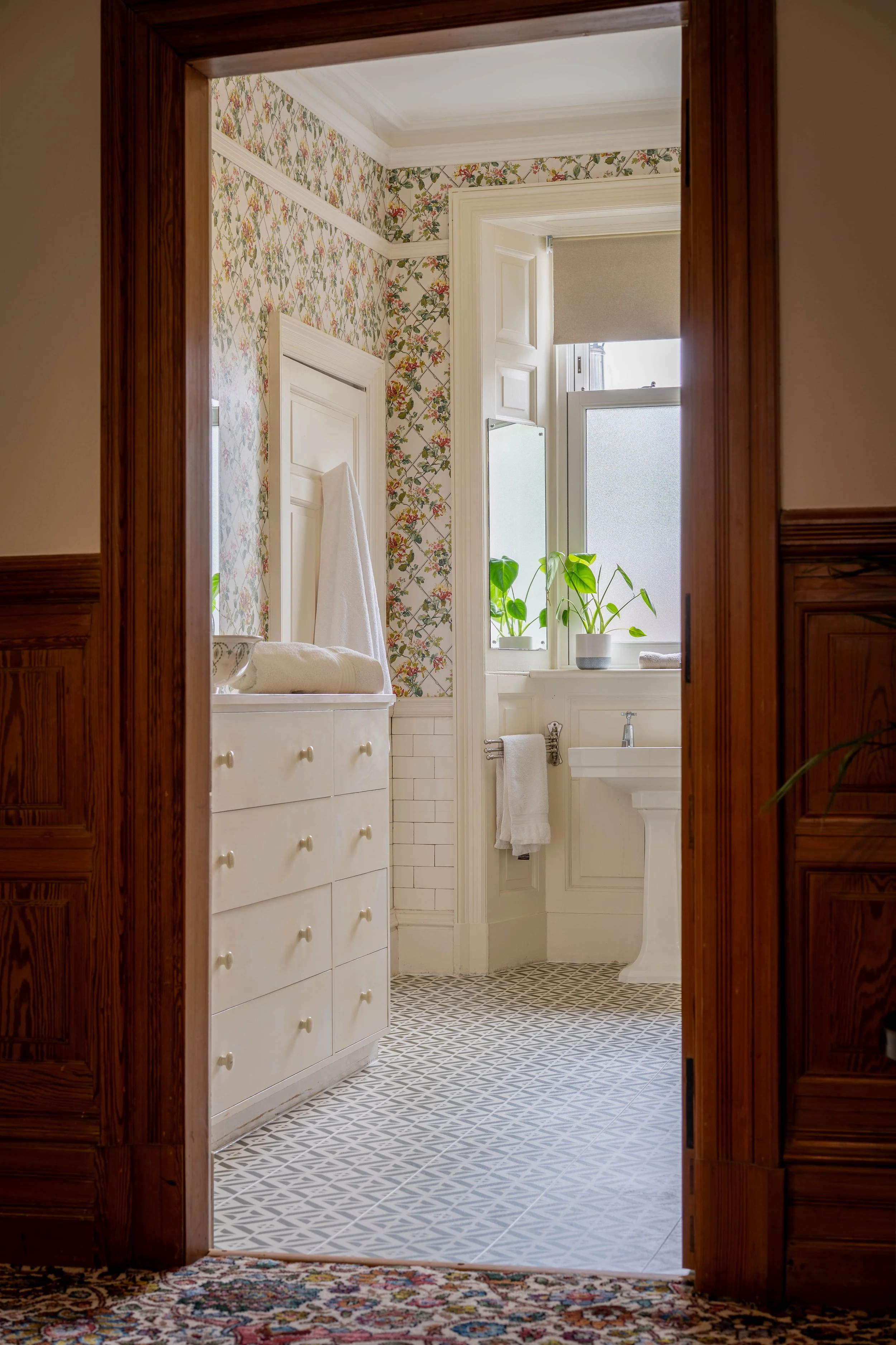 View of a vintage bathroom through a wooden doorway featuring patterned floor tiles, a white dresser with towels, a window with plants, and a pedestal sink.