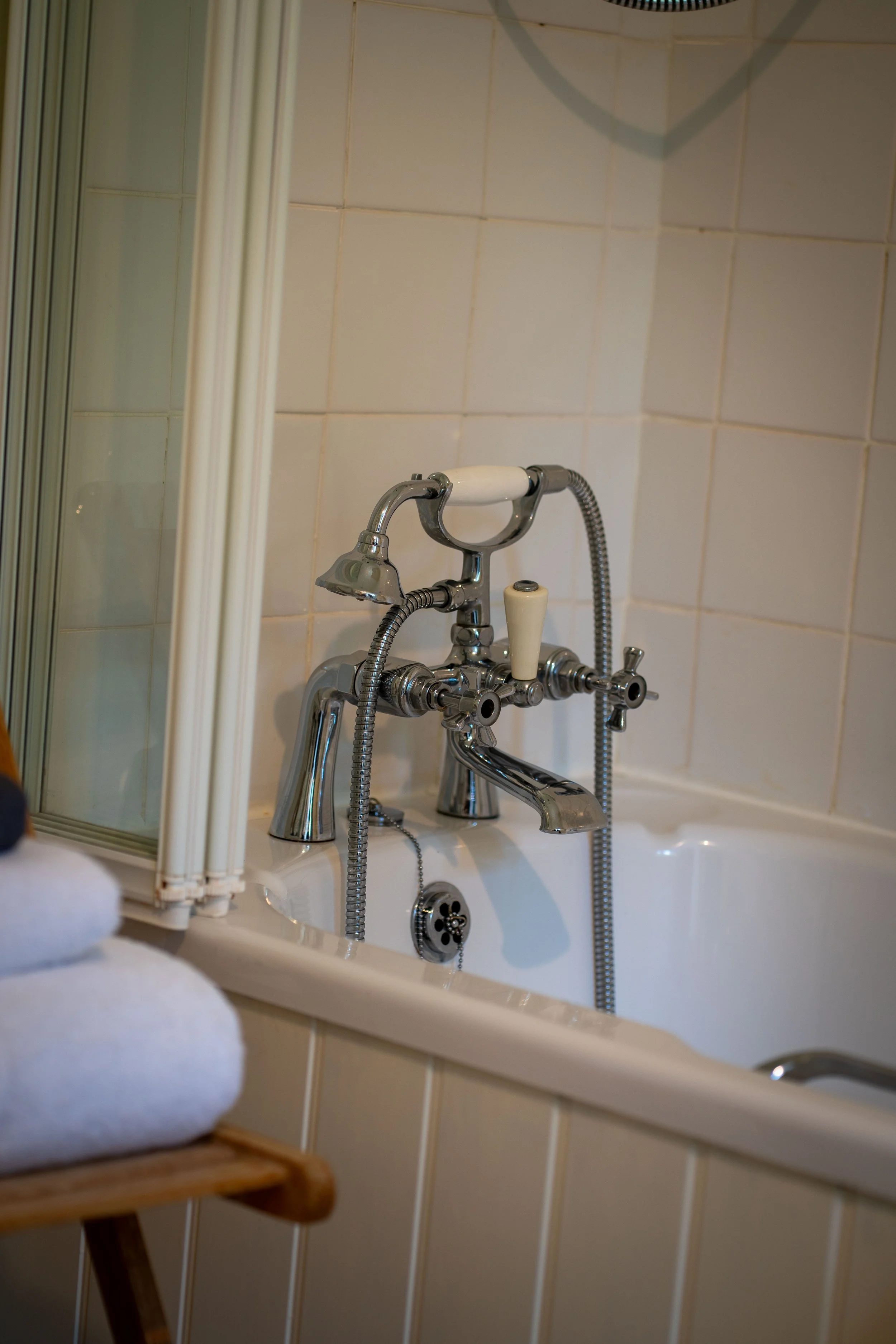 Close-up of a vintage-style bathtub faucet and handheld showerhead mounted on a tiled bathroom wall, with a reflective mirror and a wooden stool with white towels in the foreground.