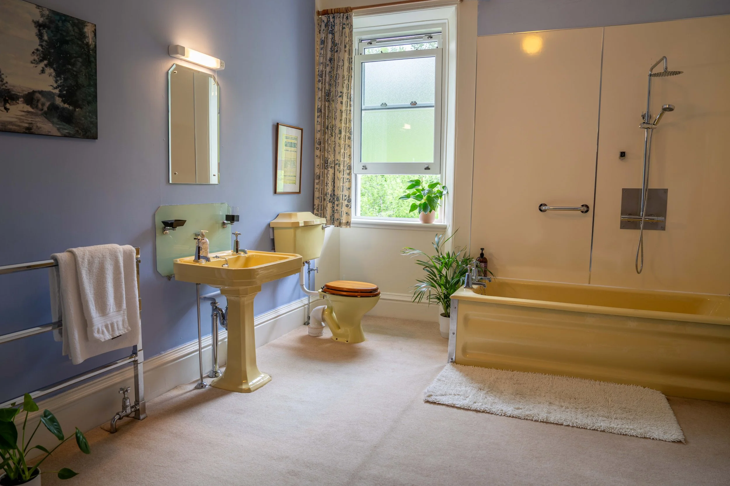 A vintage bathroom with pale yellow fixtures, including a sink and toilet, a yellow bathtub, and a large window with floral curtains. The room has light blue walls, framed pictures, a mirror, a towel rack, and potted plants, with sunlight coming thro