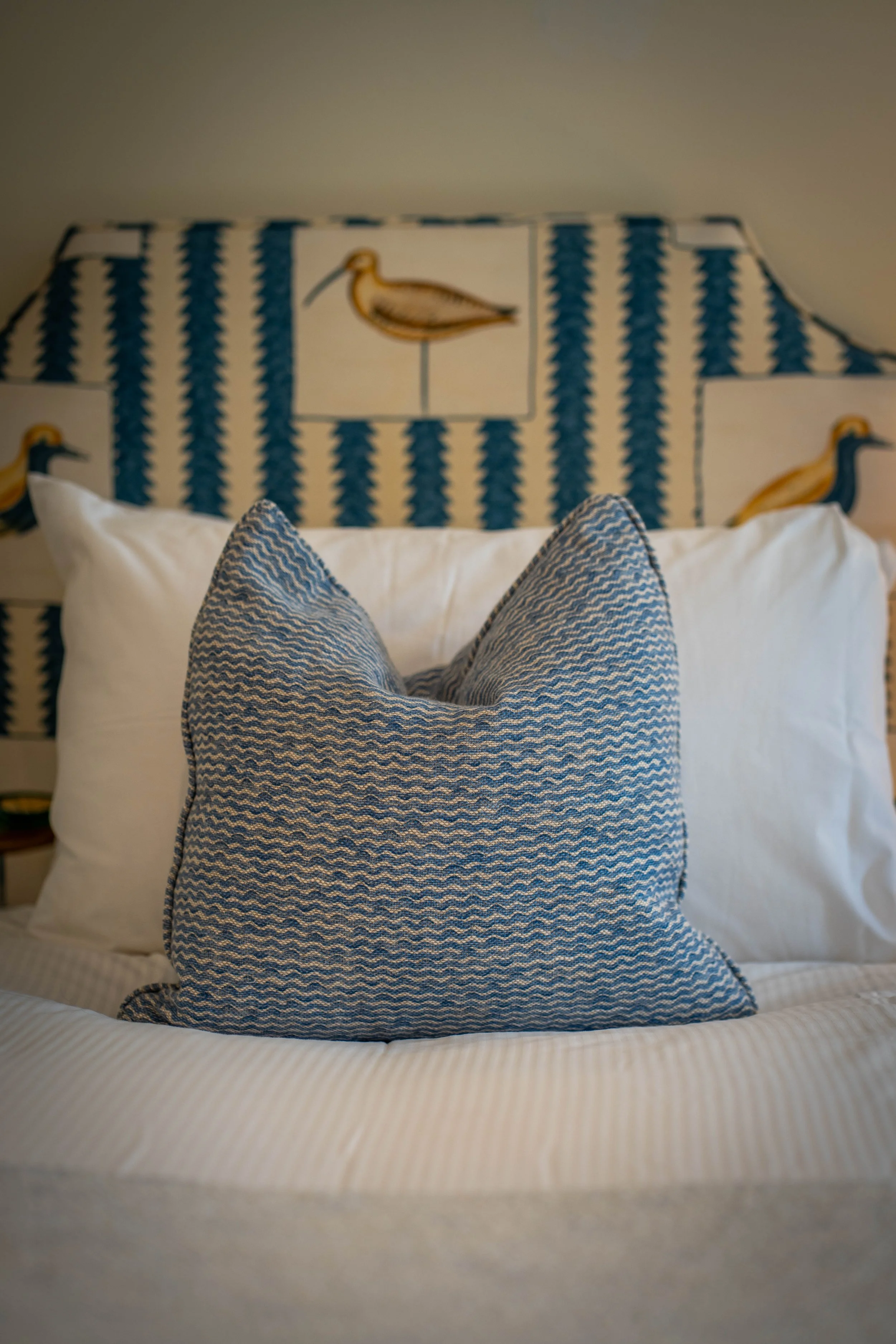 A close-up view of a decorative pillow on a bed with a chevron pattern and a bedspread with a similar zigzag pattern in a bedroom with a patterned headboard featuring birds and stripes.