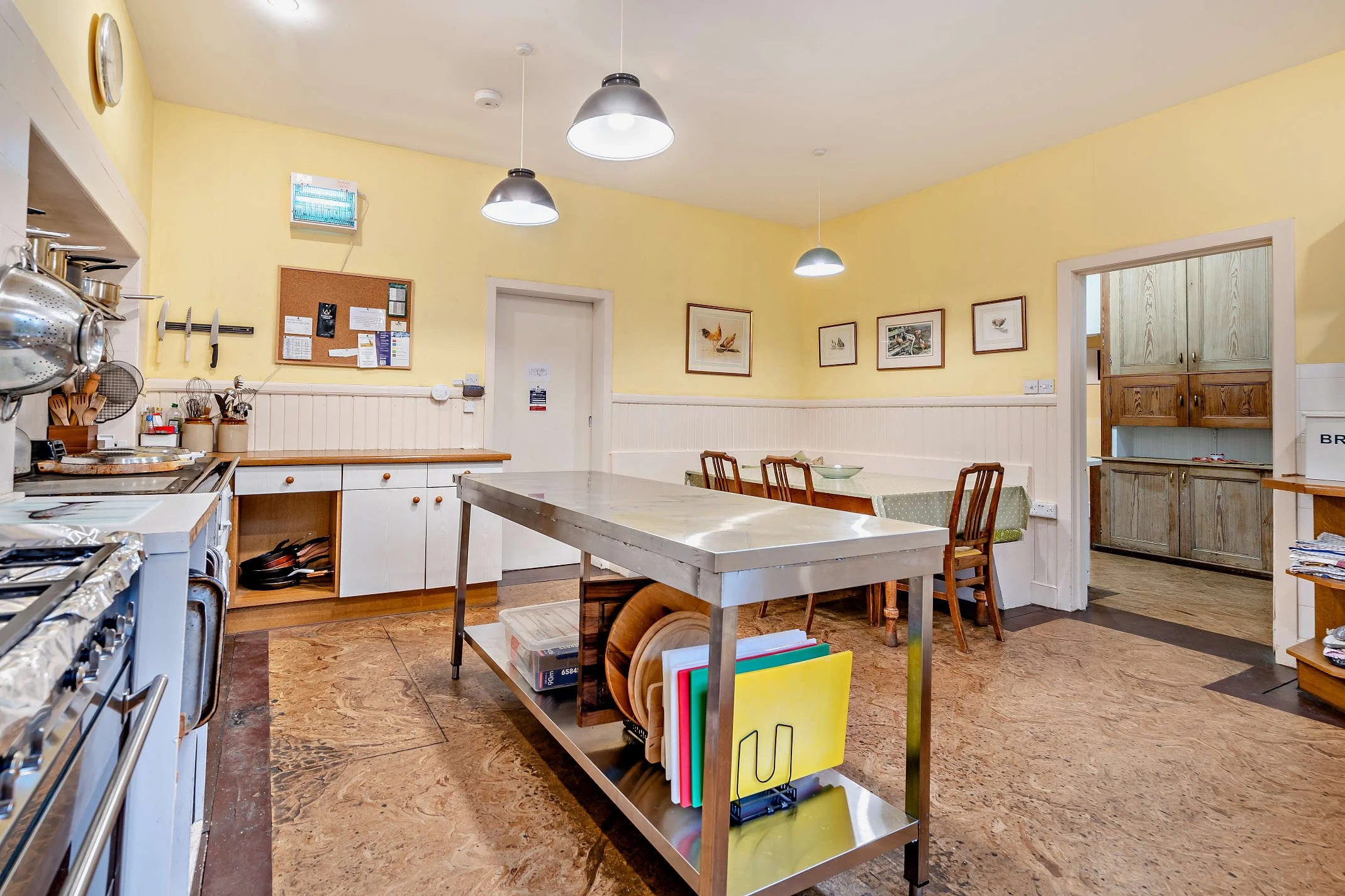 A kitchen with yellow walls and white cabinets, featuring a metal island with file folders underneath, a wall clock, framed pictures of birds, and various kitchen utensils and appliances visible on the countertops and hanging racks.