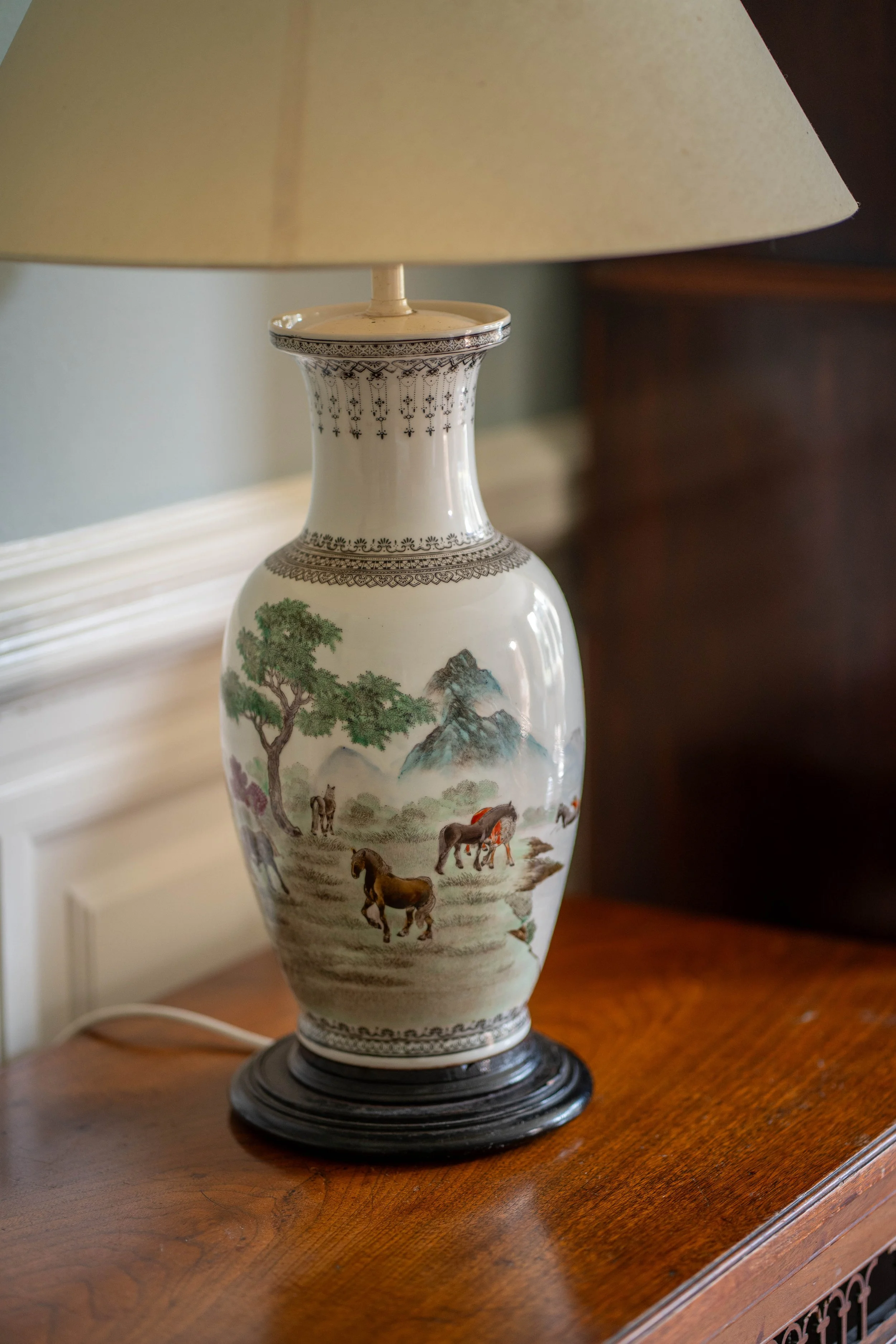 A decorative porcelain table lamp with traditional landscape scene and horses, sitting on a wooden surface.