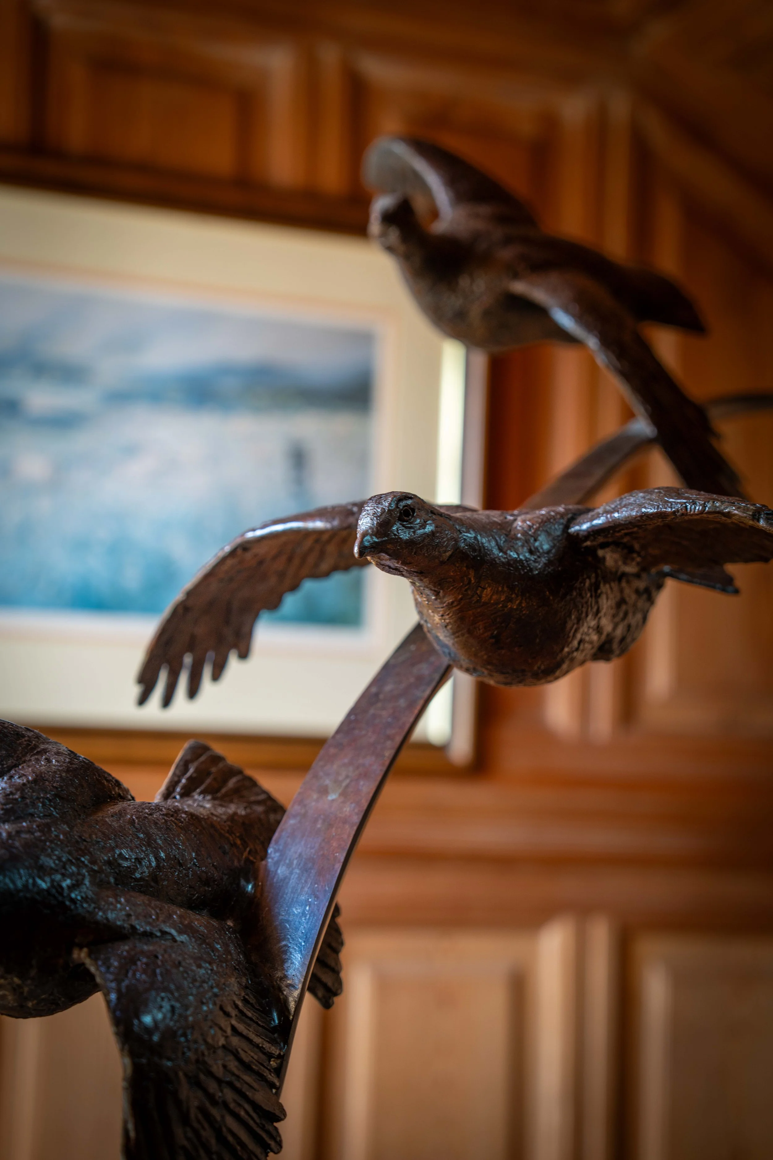Close-up of a bronze sculpture of birds in flight with a blurred background of a framed painting and wooden wall.