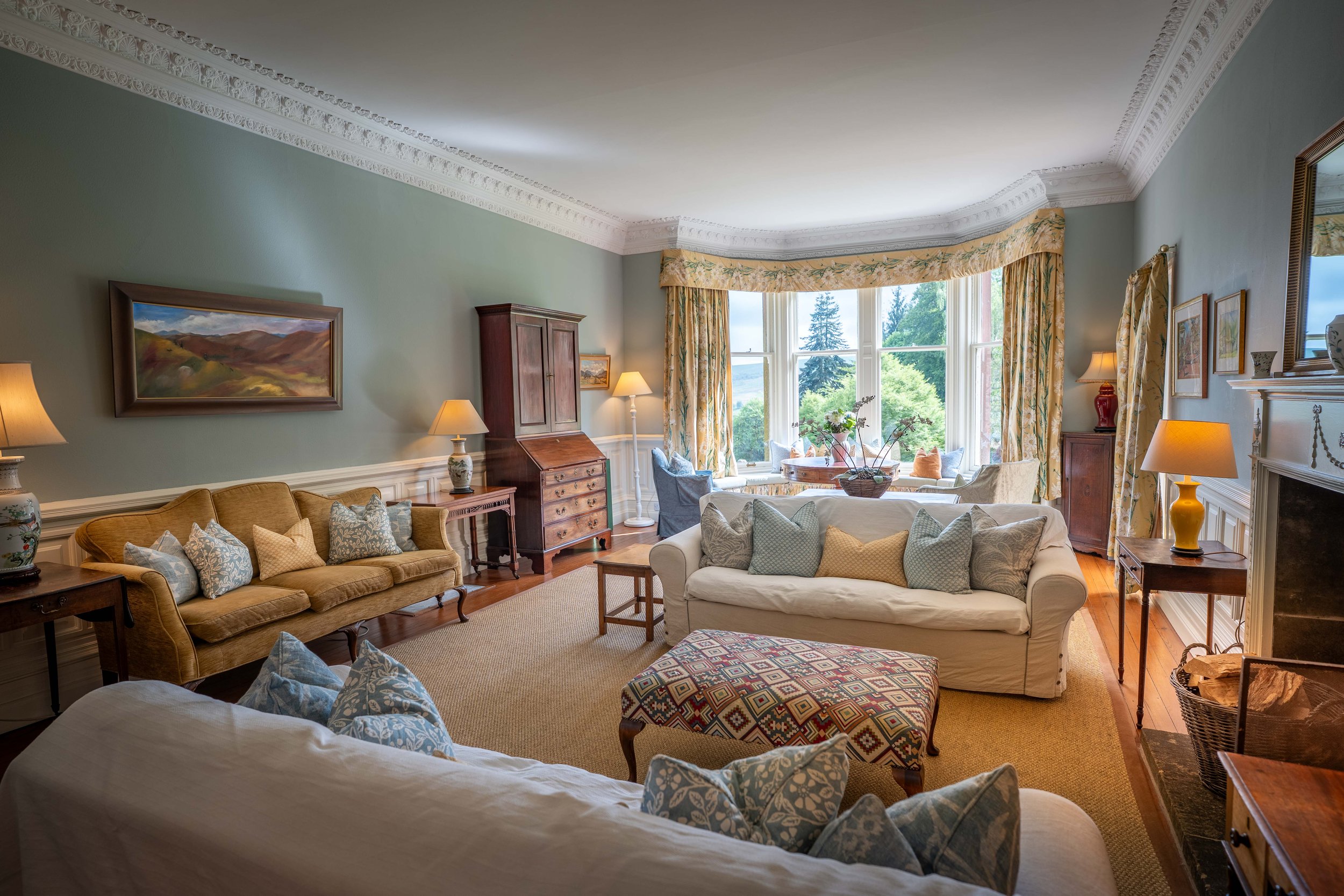 A cozy living room with two white sofas, a yellow sofa, and multiple throw pillows, situated near large bay windows with floral curtains, allowing natural light and a view of greenery outside. The room features framed paintings, lamps, a wooden cabin