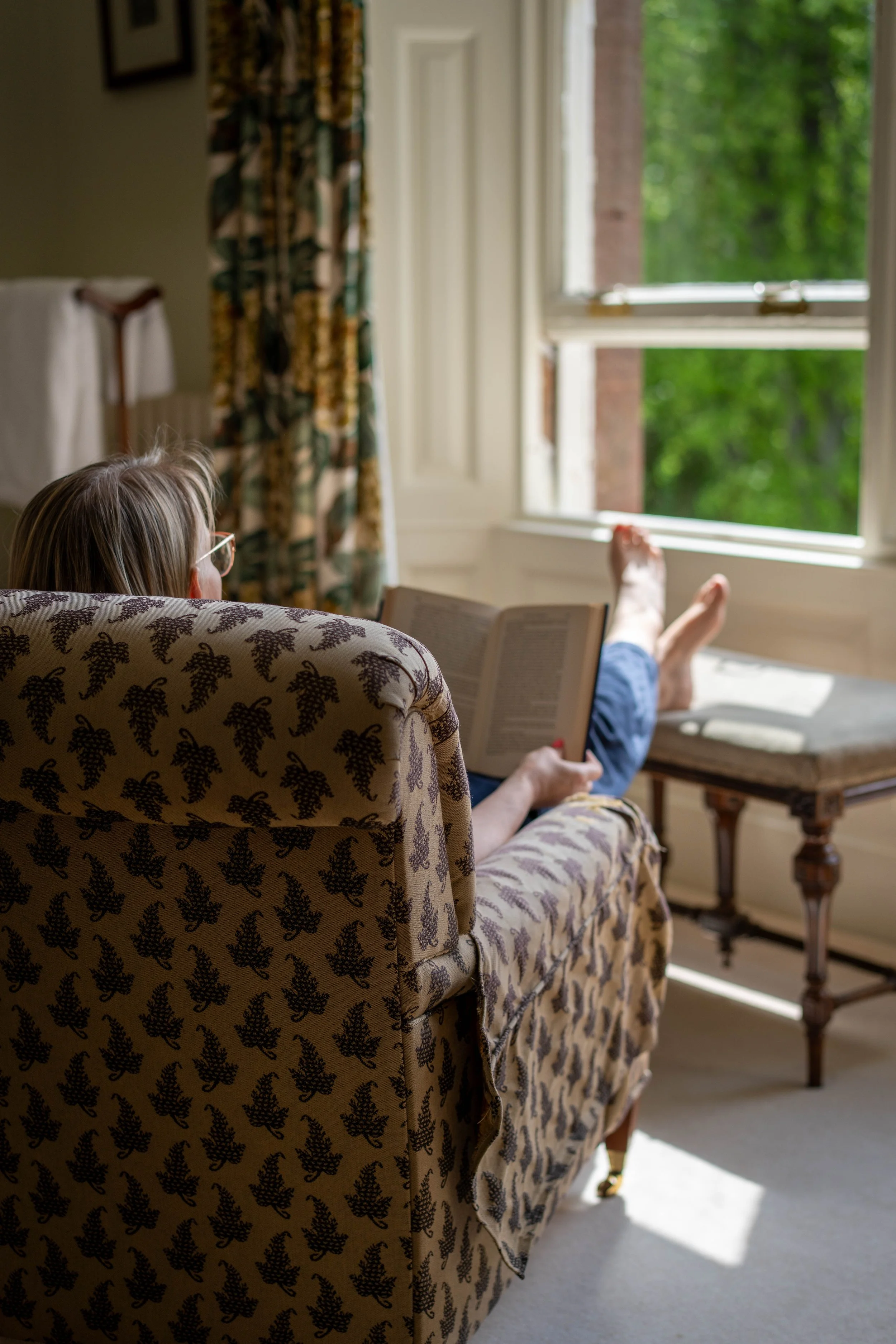 A woman with glasses sitting on a floral upholstered armchair, reading a book, with her feet up on a wooden table near a window with open shutters and green foliage outside.