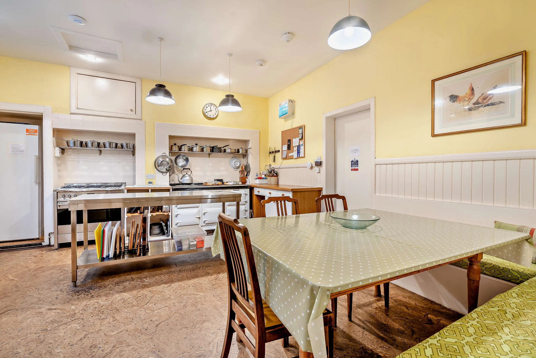 A cozy kitchen with yellow walls and a dining table with a green polka dot tablecloth, wooden chairs, and a decorative framed picture of roosters on the wall. The kitchen features hanging pendant lights, open shelving with pots and pans, and a clock 
