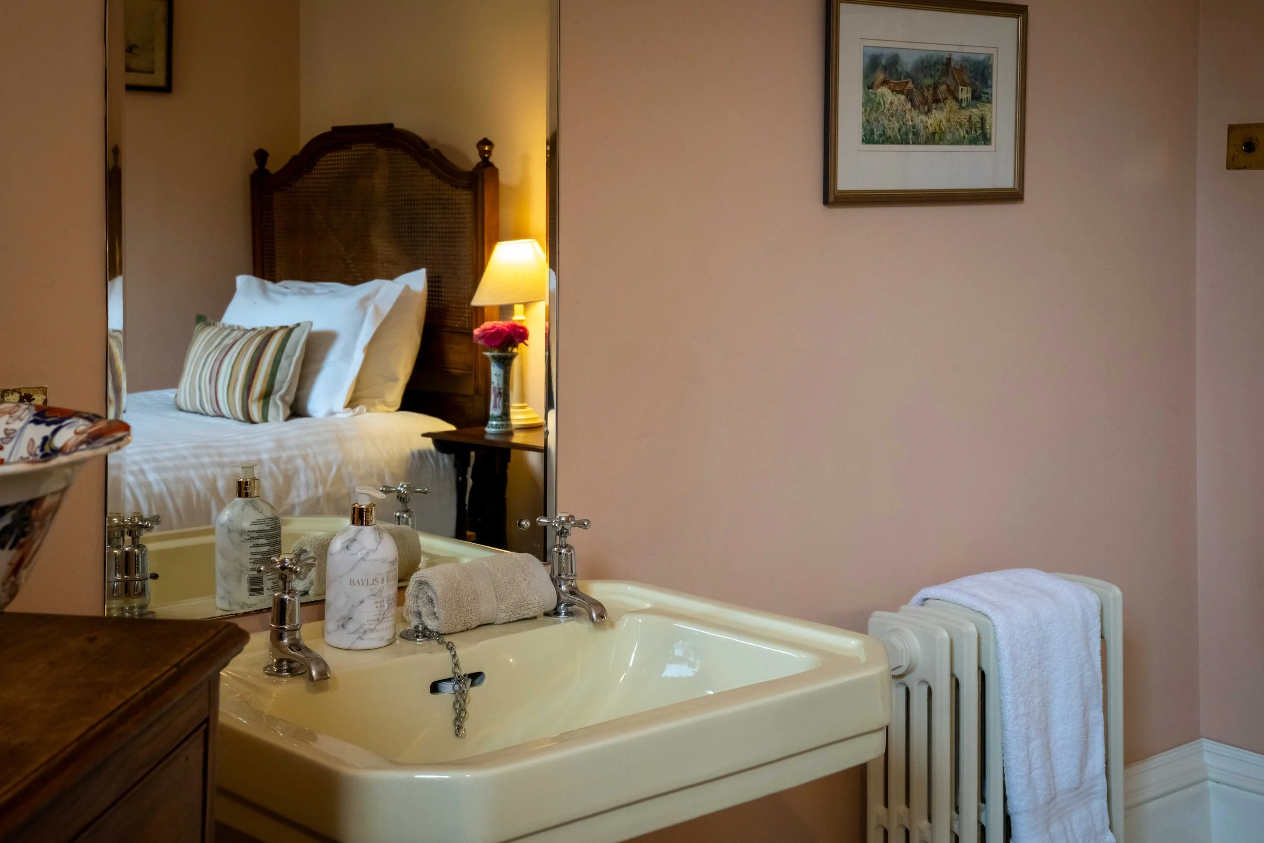A hotel room with a bed visible through a mirror reflection, a lamp, framed artwork, a nightstand, a sink with toiletries, a rolled towel, and a radiator with a towel hanging on it.