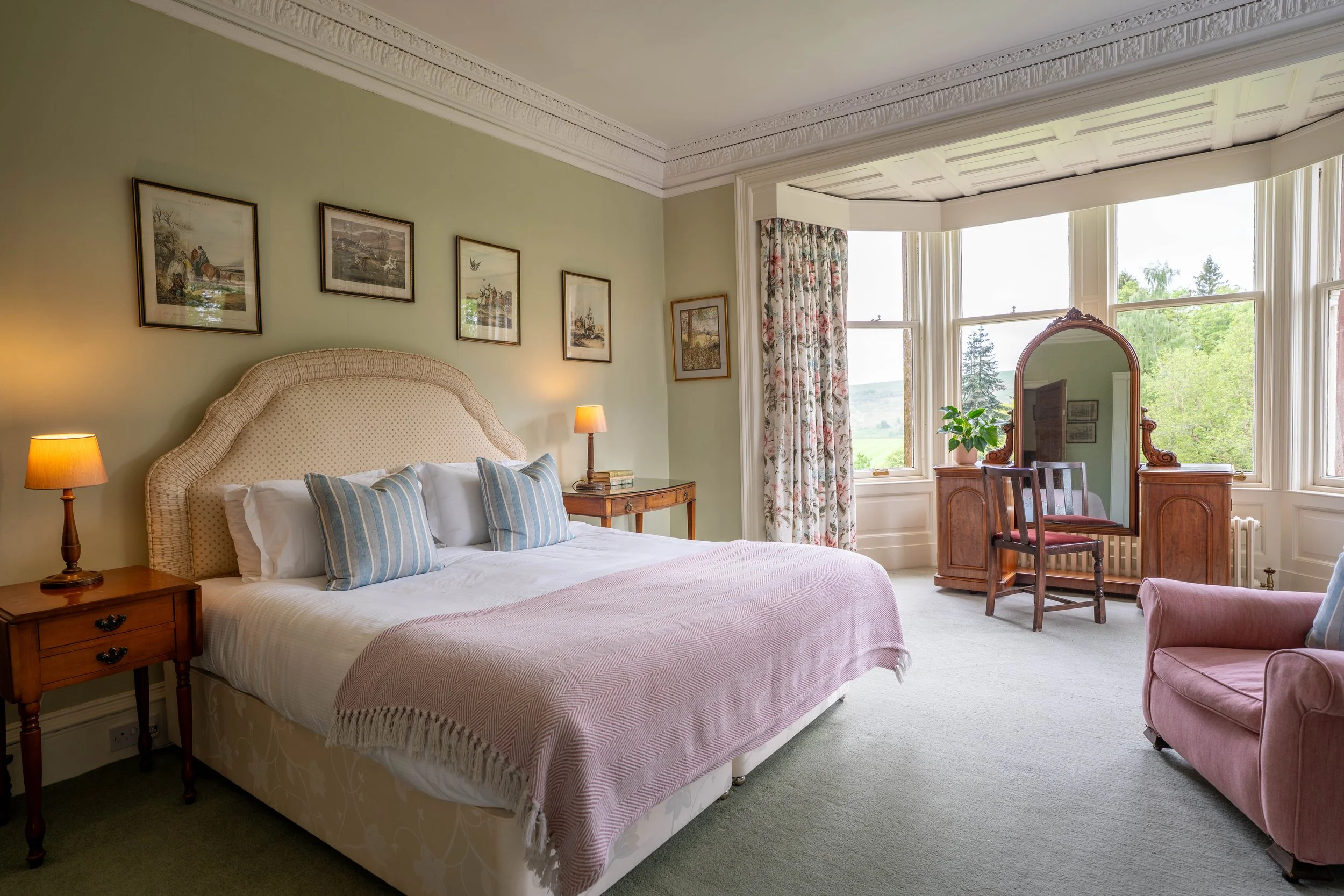 A bedroom with a large bed, striped pillows, and a bedside table with a lamp. The wall above the bed has four framed pictures. Large windows let in natural light, revealing trees and a scenic view outside. There is a vintage-style mirror, a wooden ch