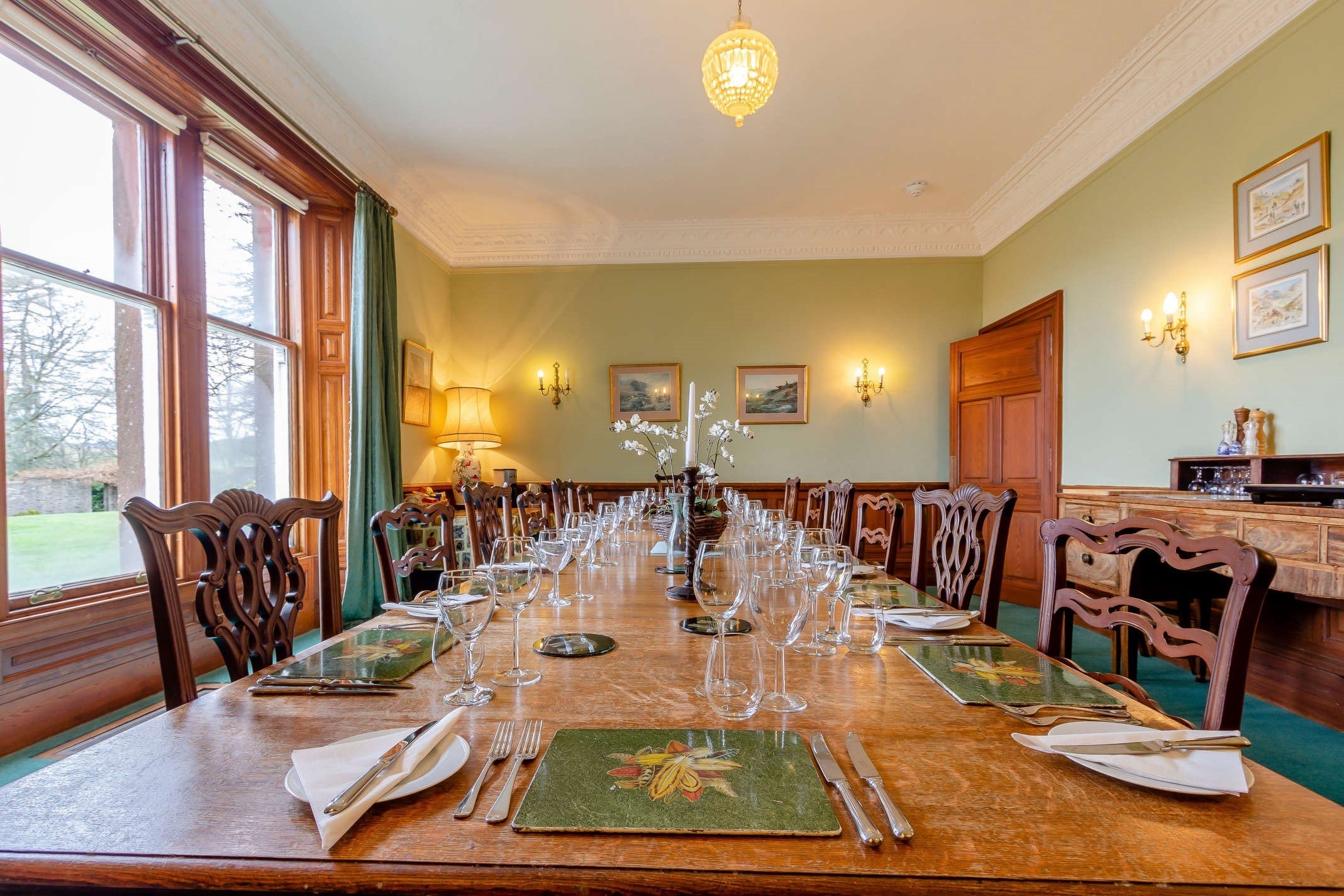 A formal dining room with a long wooden table set with glassware, napkins, and placemats, surrounded by wooden chairs, with a large window, green curtains, and framed artwork on light green walls.