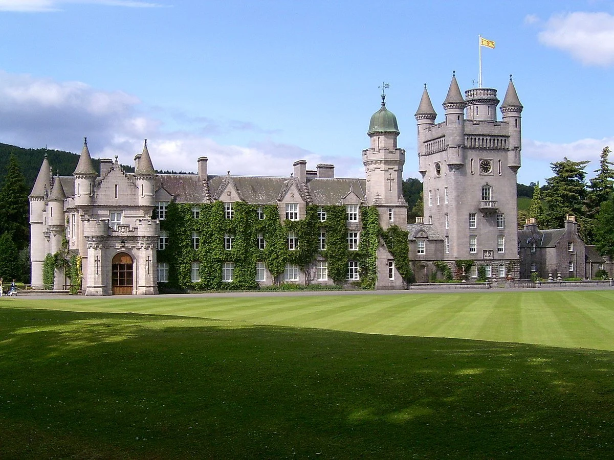 A large historic castle with multiple turrets and a flag on top, surrounded by a well-maintained grassy area and trees in the background.