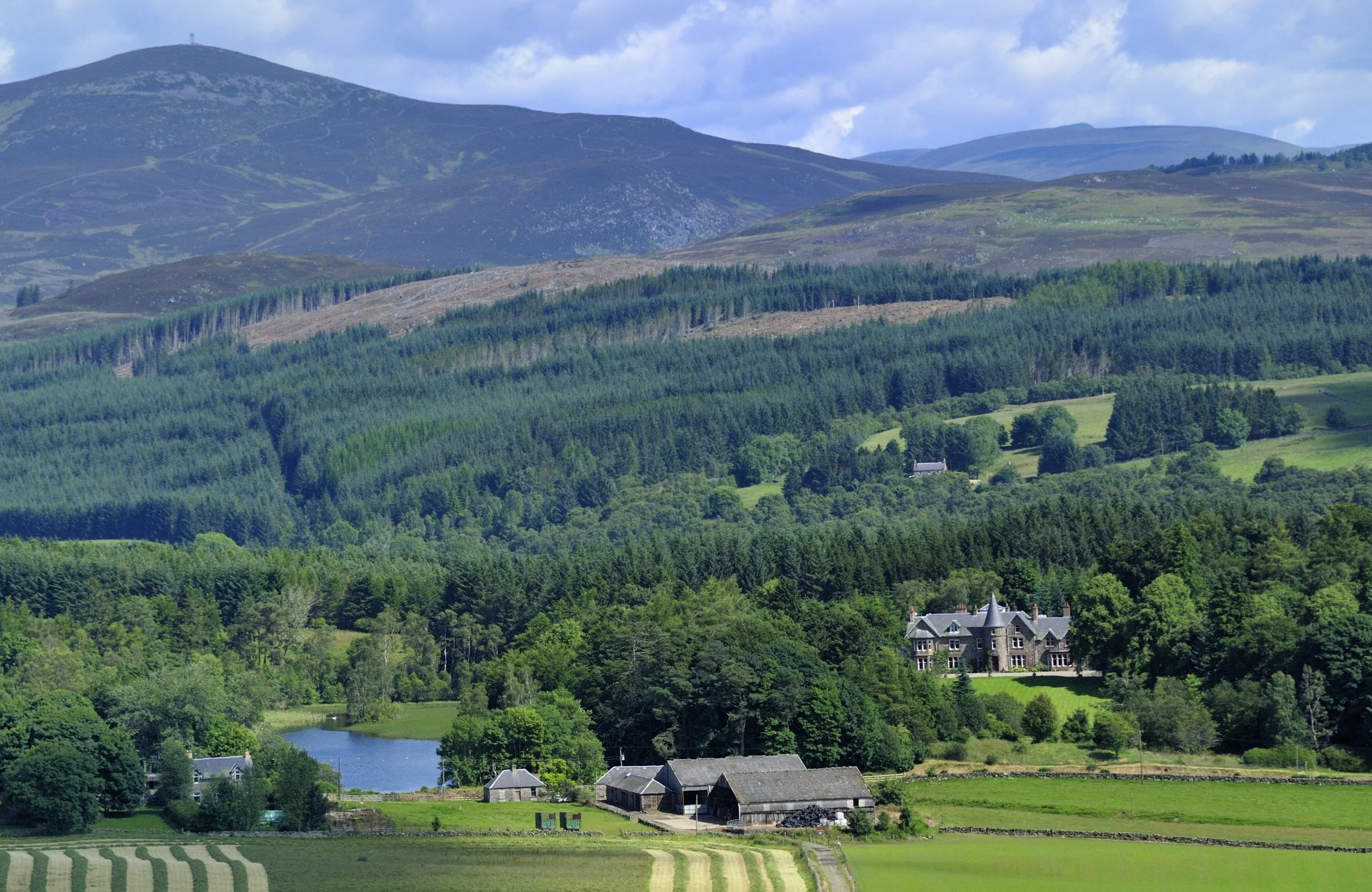 A scenic landscape of rolling green hills, dense forests, a small lake, and a large castle-like house in the foreground, with mountains in the background under a partly cloudy sky.