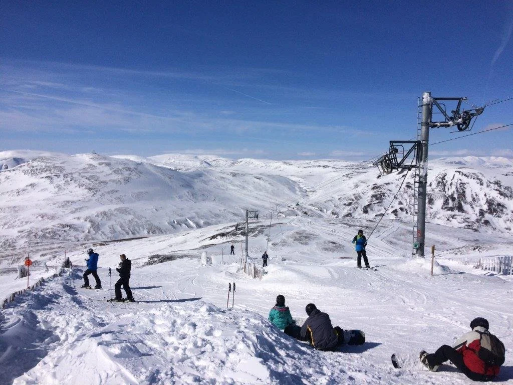 People on a snowy mountain ski slope with ski lifts and snow-covered hills in the background under a clear blue sky.