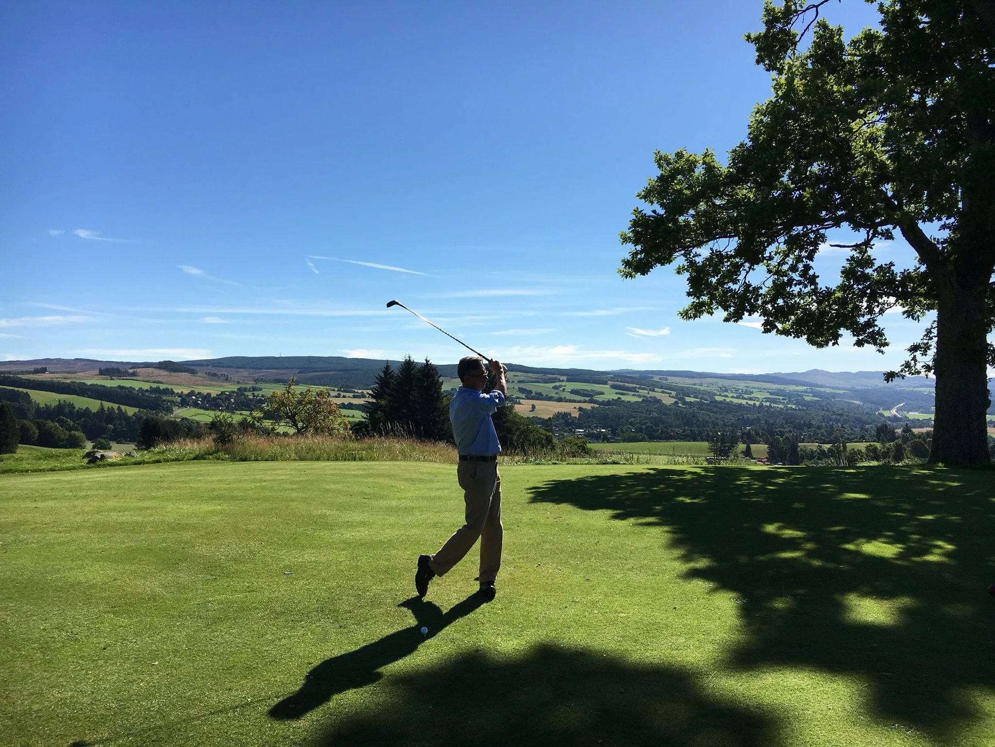 A man in a blue shirt and beige pants playing golf, swinging his golf club on a sunny day at a golf course with trees and a landscape of rolling hills in the background.