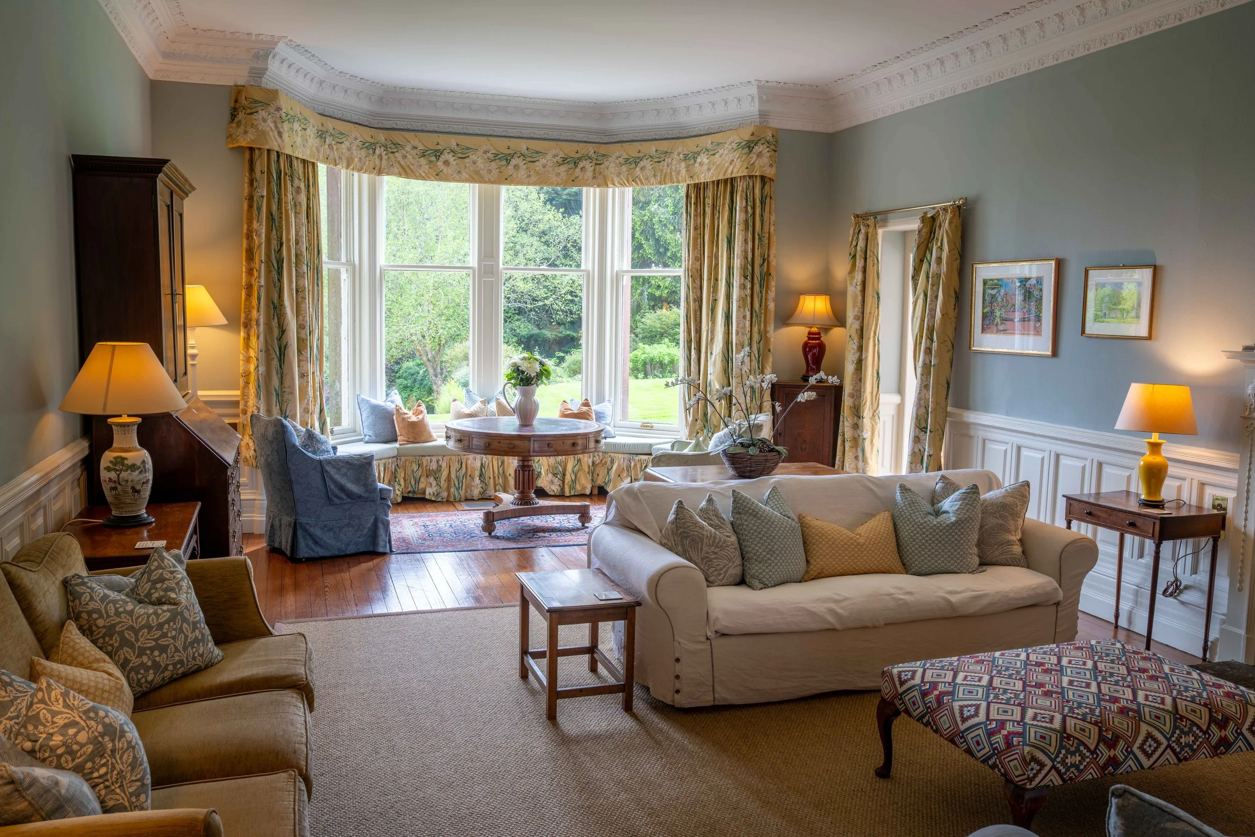 Living room with large bay window, beige sofa, armchairs, colorful cushions, wooden furniture, and framed pictures on the walls.