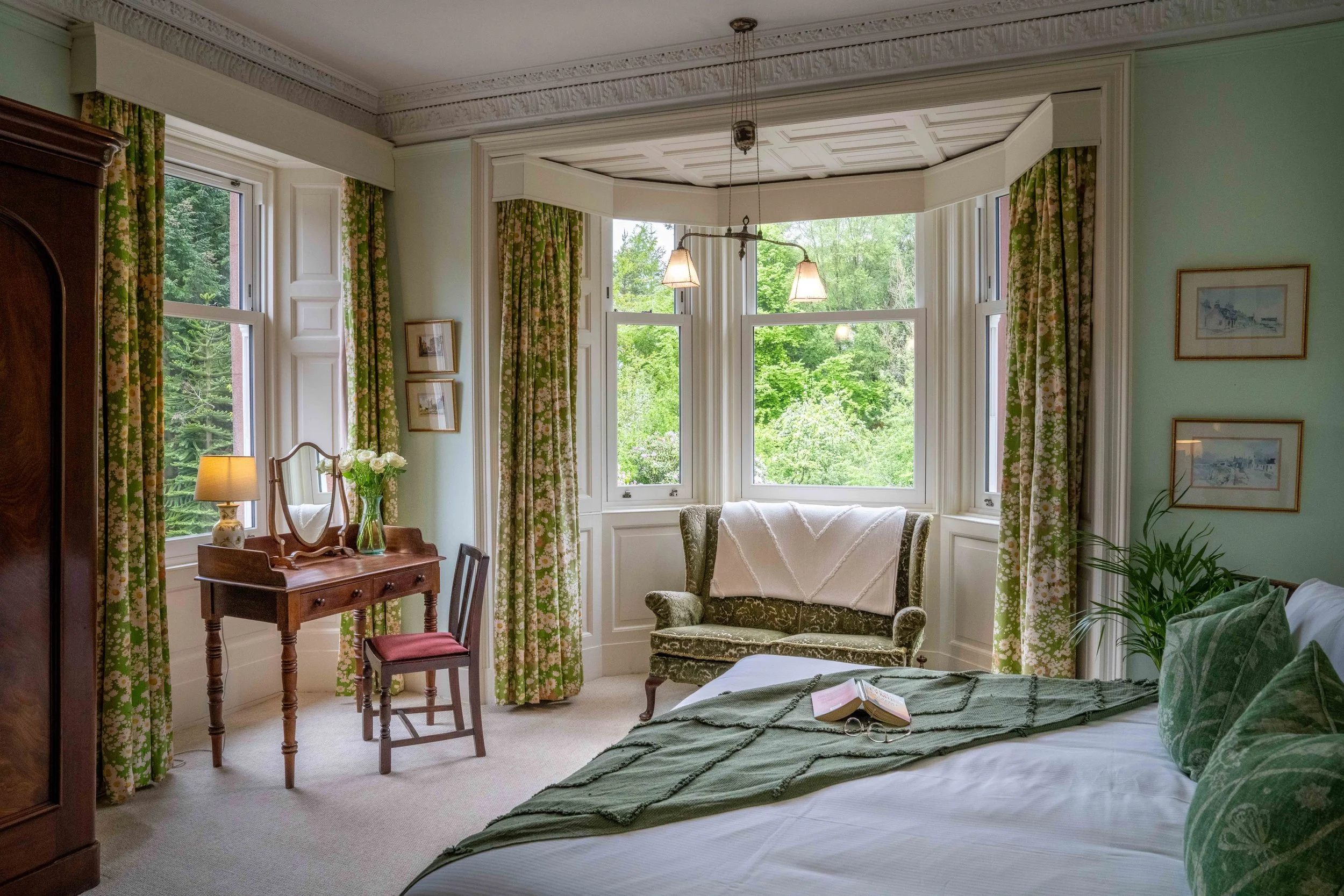 A cozy, well-lit bedroom with large windows and green, floral-patterned curtains. There is a vintage armchair, a small table with a vase of white roses, and framed pictures on the wall. The bed has a green quilt and an open book with reading glasses on top, with greenery visible outside the windows.