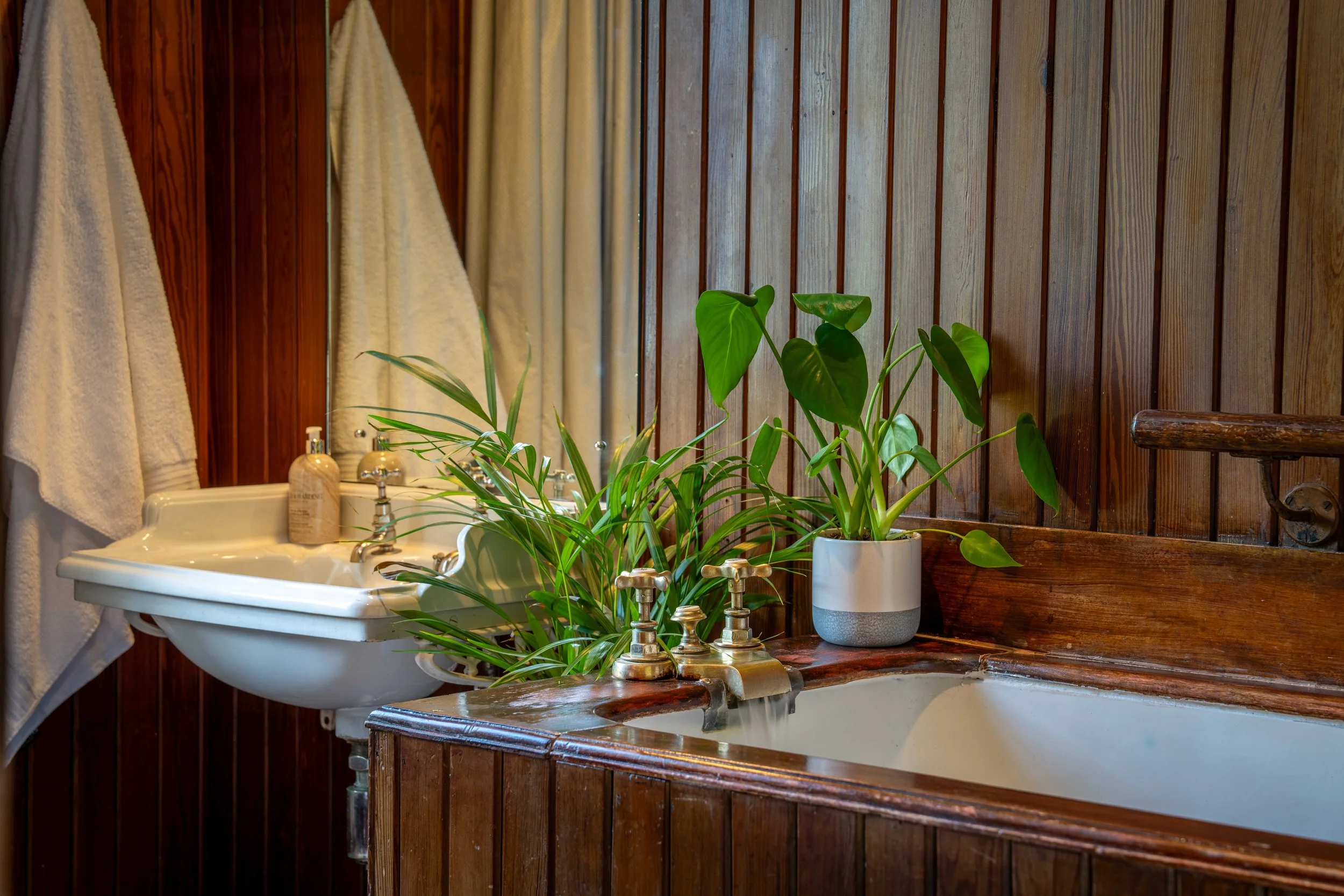 A bathroom with wooden wall paneling and a white sink, decorated with several green potted plants, towels hanging on hooks, and bottles on the sink.