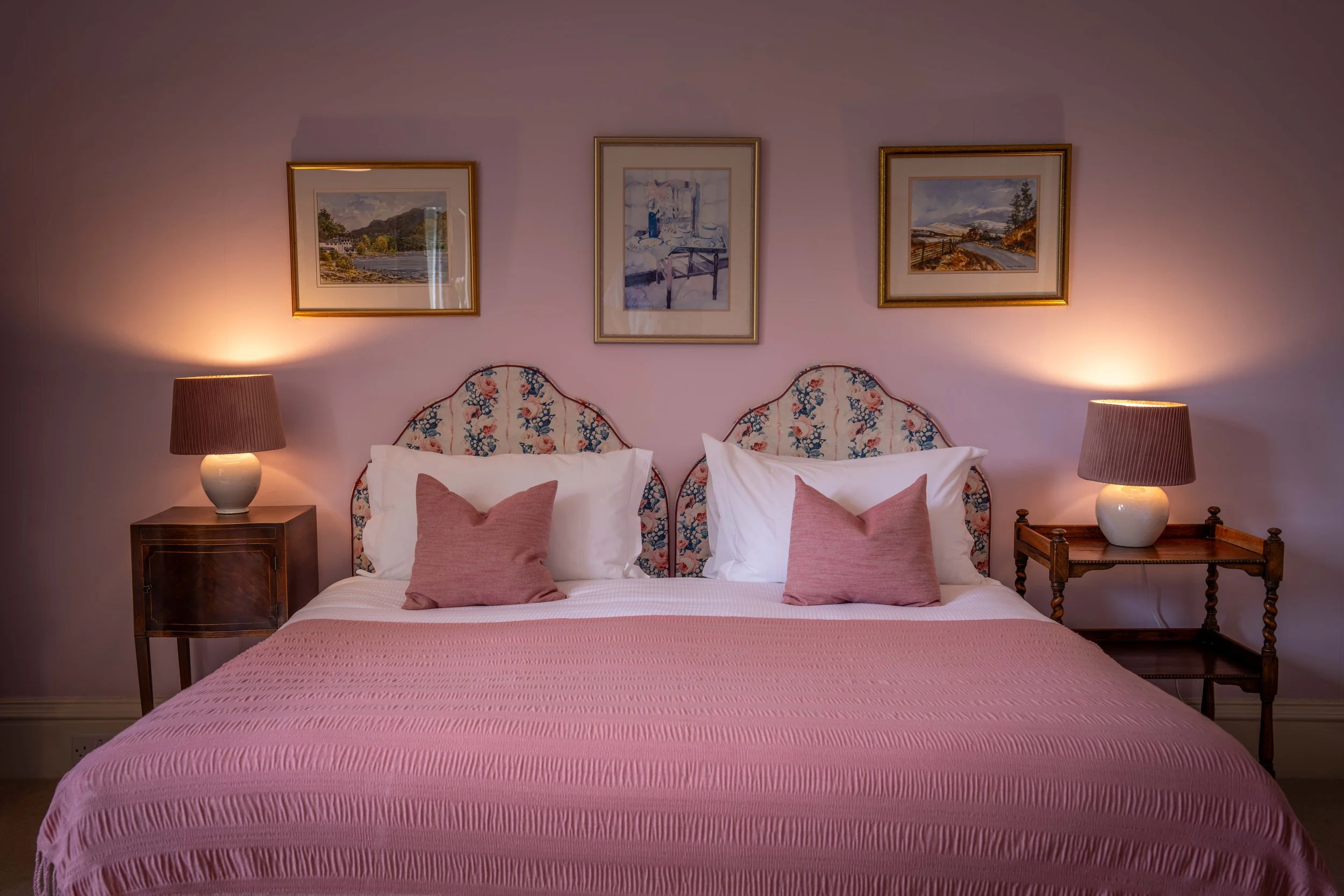 A bedroom with two twin beds, pink bedspreads, and matching pink pillows, flanked by two wooden nightstands with table lamps. Above the beds are three landscape paintings in gold frames.