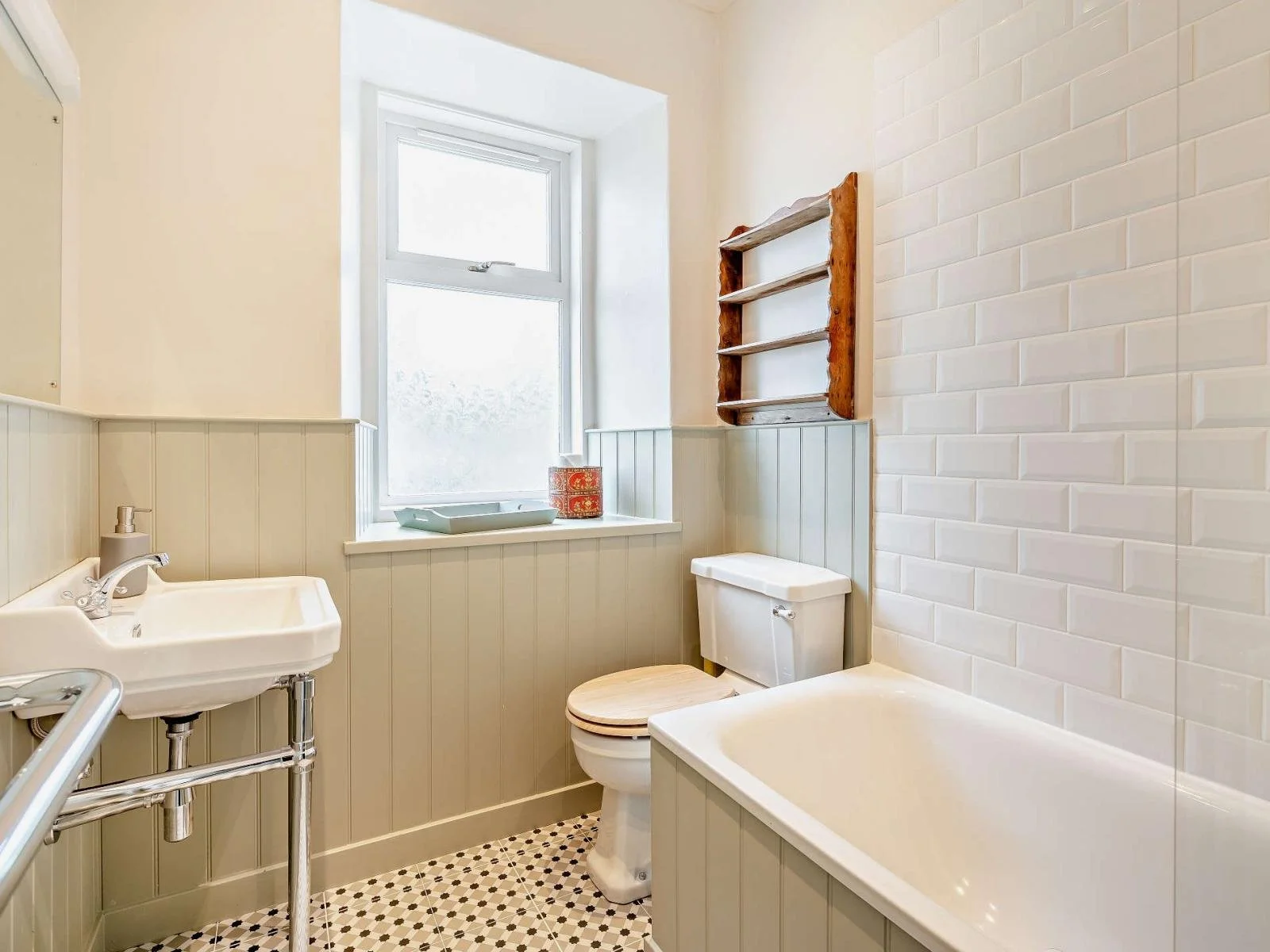 Interior of a small bathroom with a bathtub, toilet, sink, and window, featuring beige wainscoting and white tile wall.
