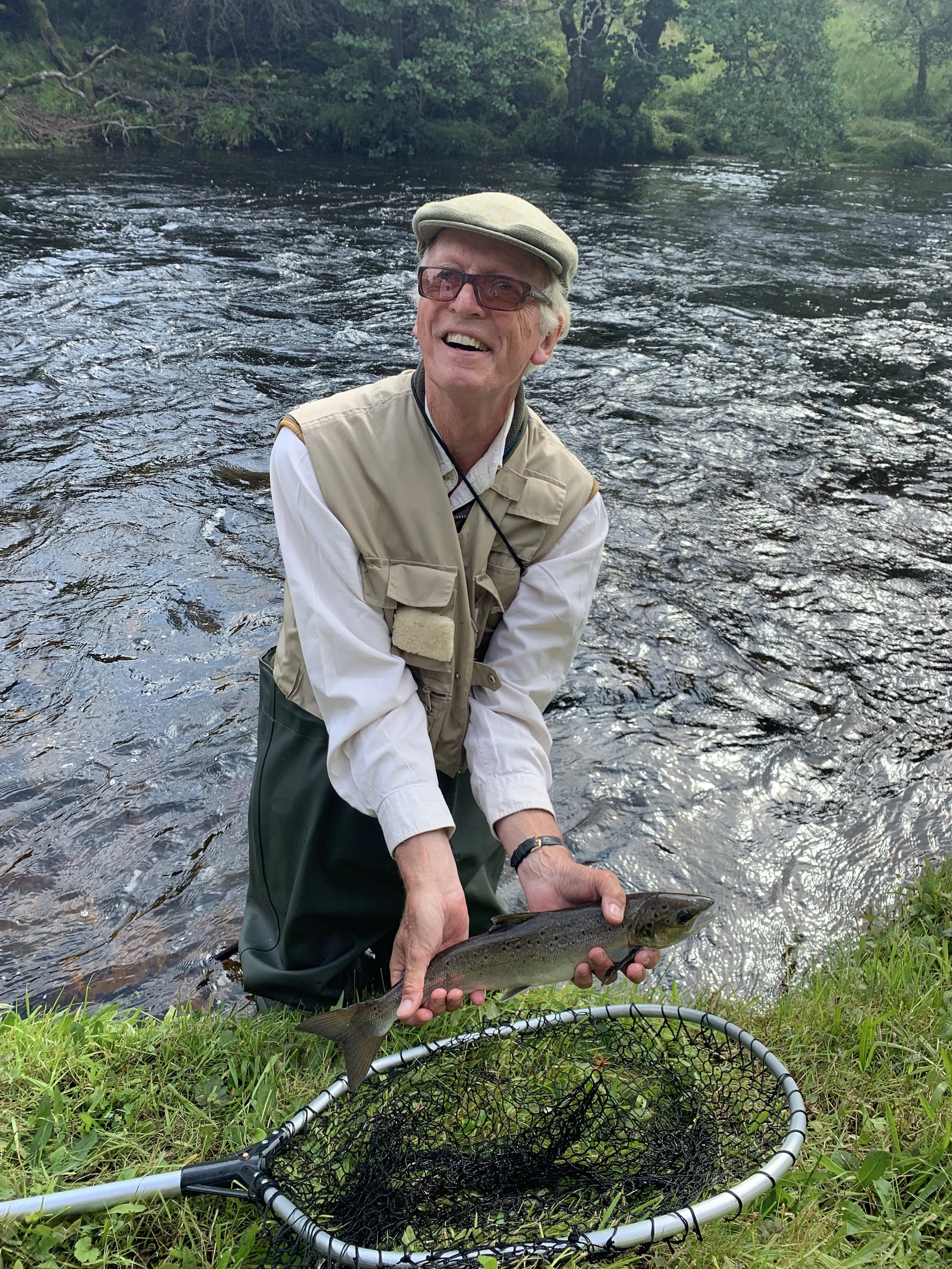 A smiling elderly man wearing a hat, glasses, and fishing gear, standing in a river, holding a caught fish with a fishing net nearby on the grass.