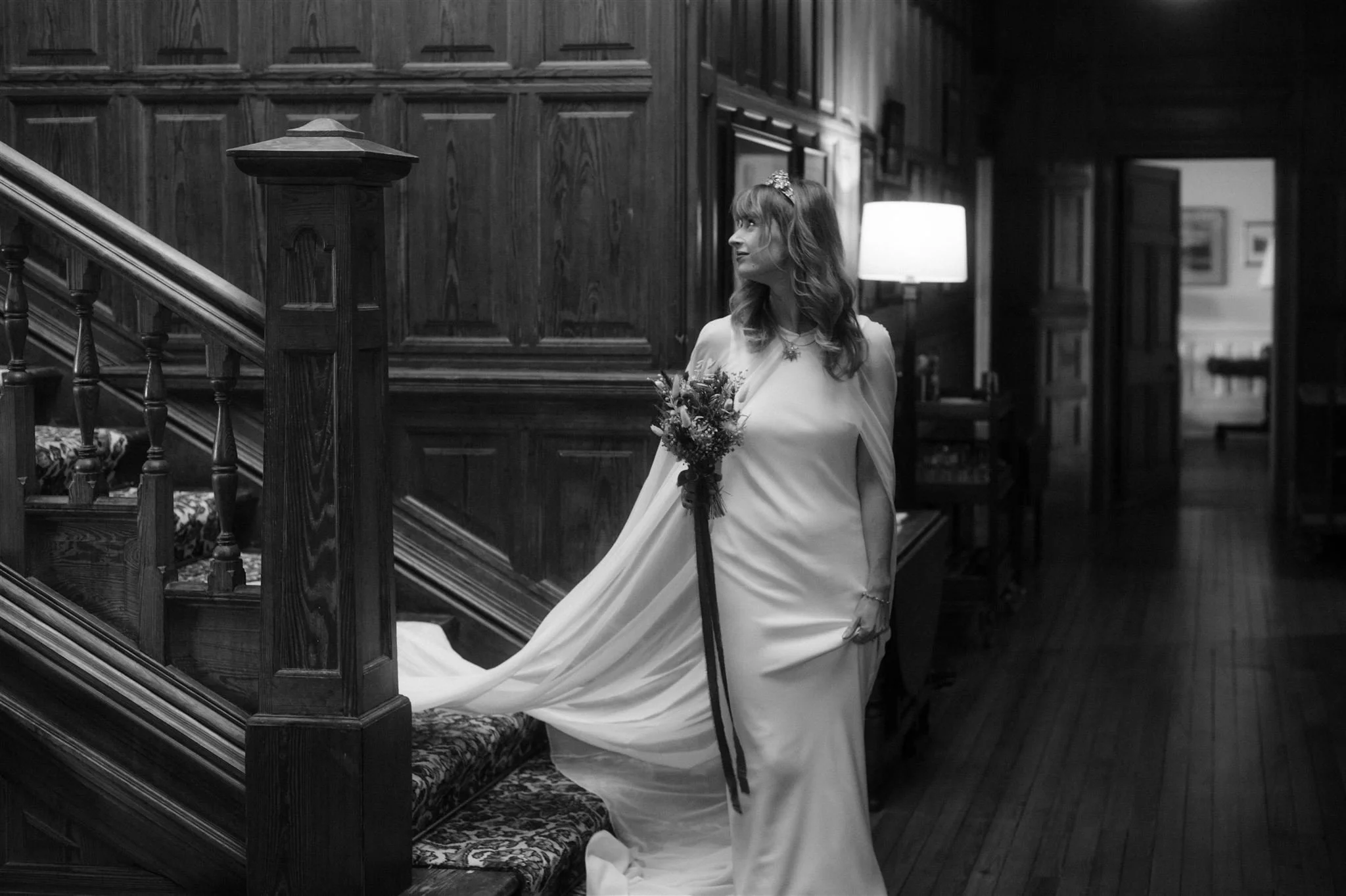 A woman in a white dress holding a bouquet of flowers standing in an elegant library with wooden paneling and a staircase.