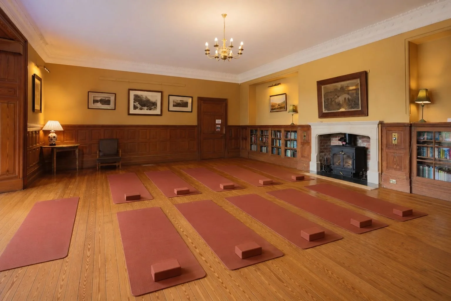 A yoga or meditation room with wooden floors, beige walls, and a fireplace. There are eight yoga mats with bricks on top arranged in two rows. The room has framed artwork, a chandelier, lamps, and built-in bookshelves.