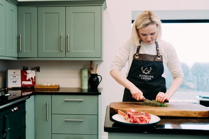 A woman preparing herbs on a cutting board in a kitchen with green cabinets, a plate of raw steak, and a window in the background.