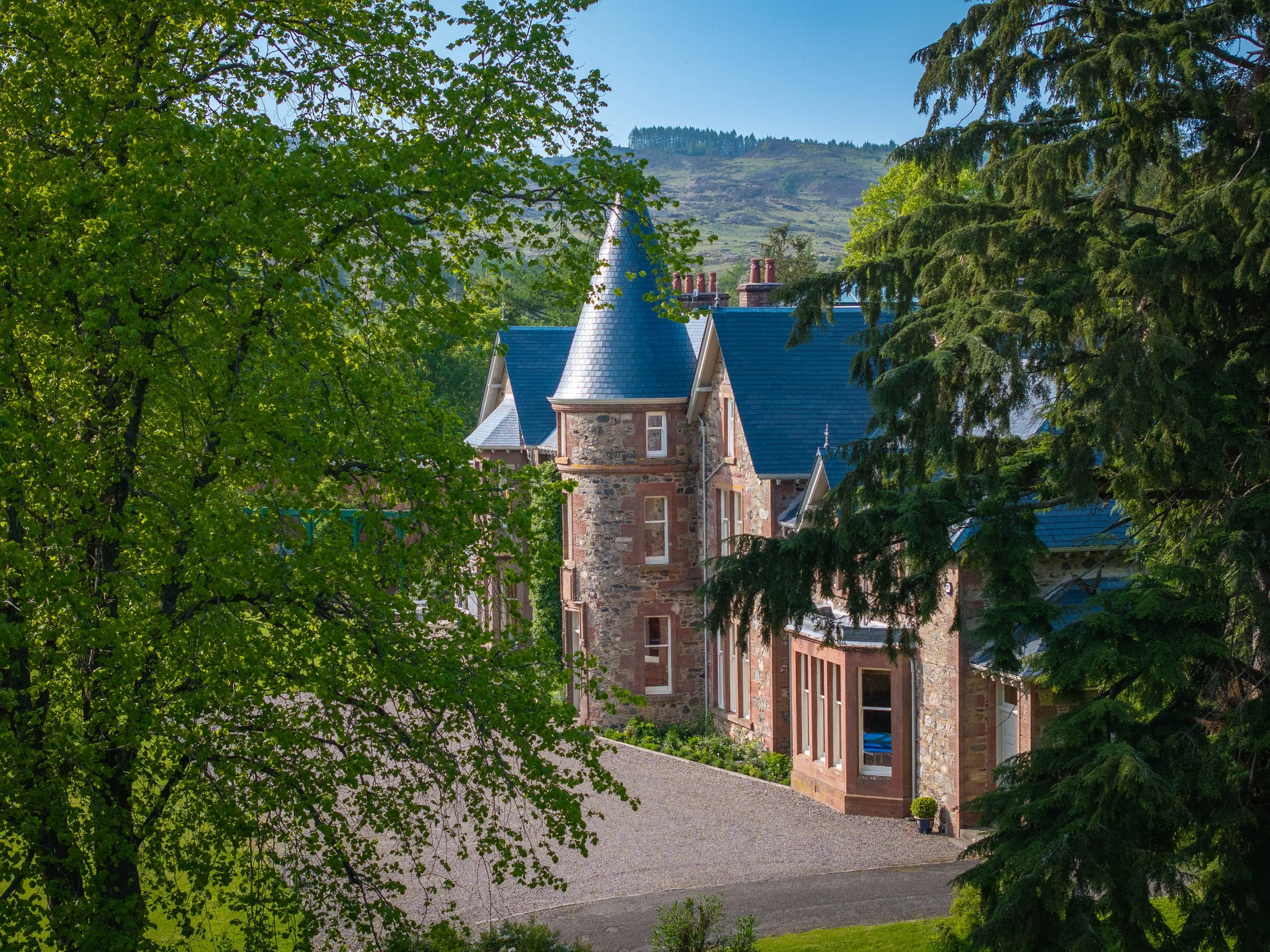 A house with turret-style towers and a blue roof is surrounded by green trees in a rural setting, with a mountain and a clear blue sky in the background.