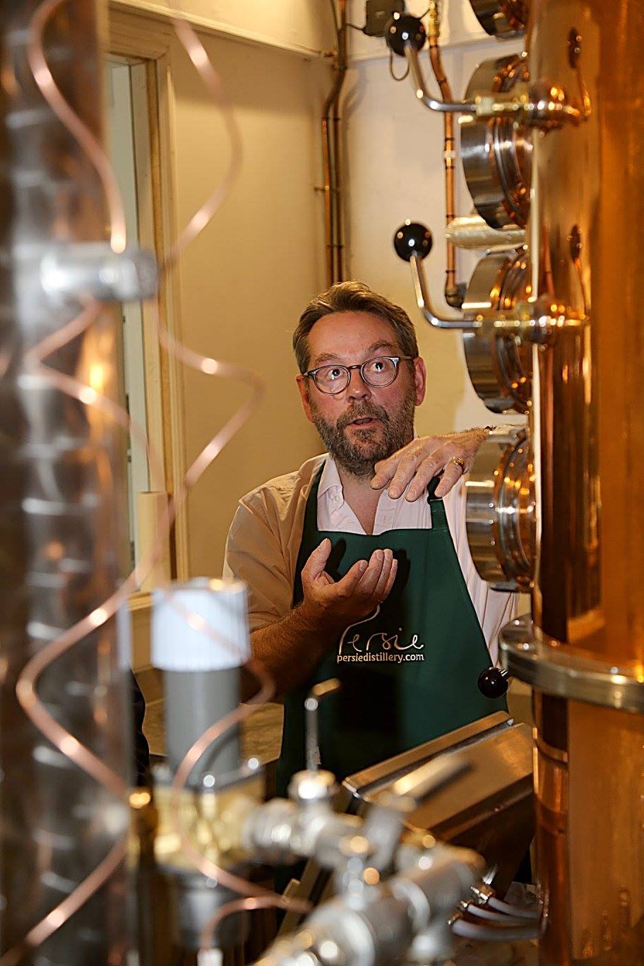 A man with glasses and a beard, wearing a green apron, stands behind a copper distillation apparatus, looking through it with a surprised expression.