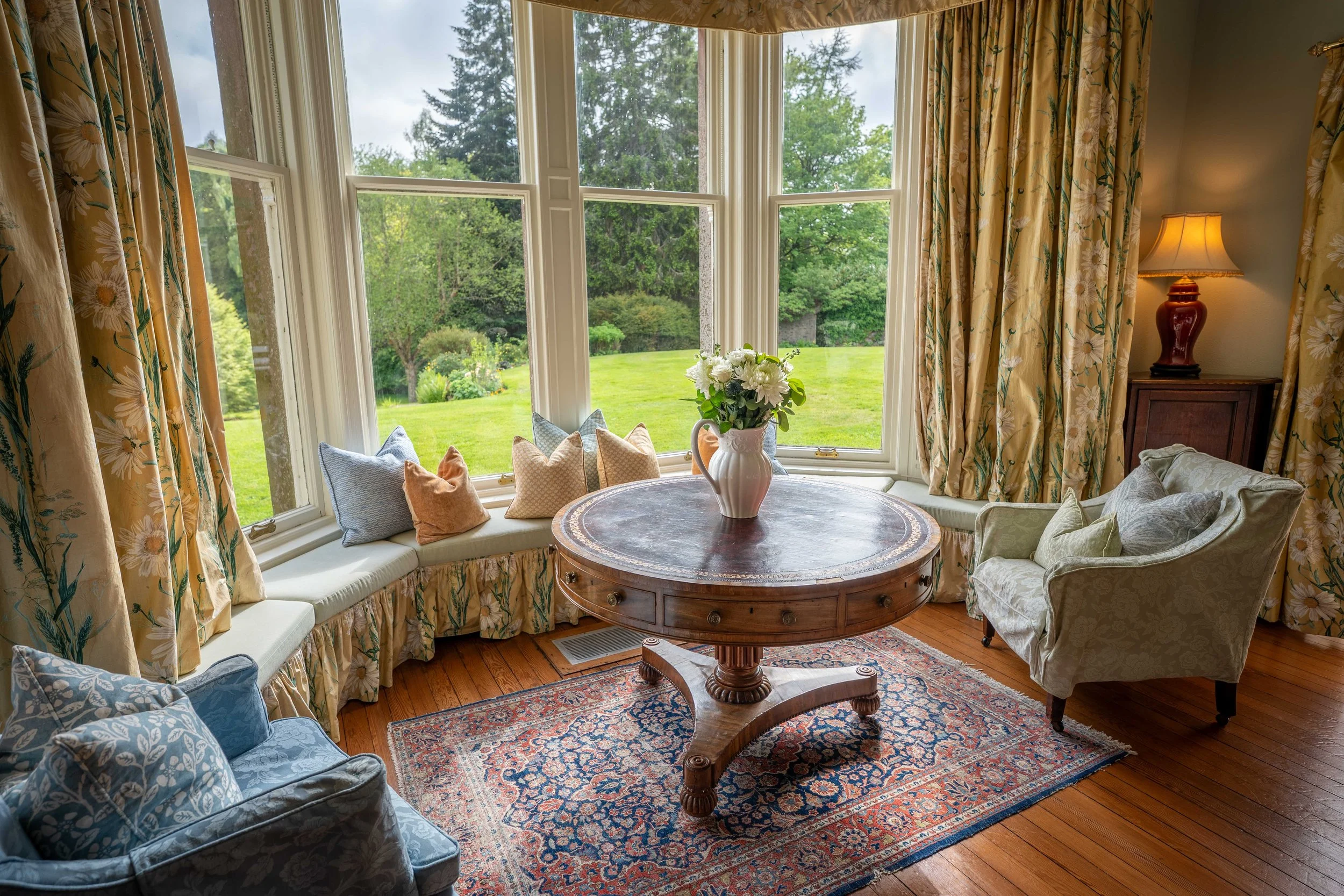 A cozy sitting area with a large bay window overlooking a green yard and trees. The window has floral curtains, and a round wooden table with a floral centerpiece is placed in front of the window. There are cushions on a built-in window seat and upholstered chairs around the table. A table lamp with a red base and beige shade provides warm lighting. An oriental rug is on the wooden floor.