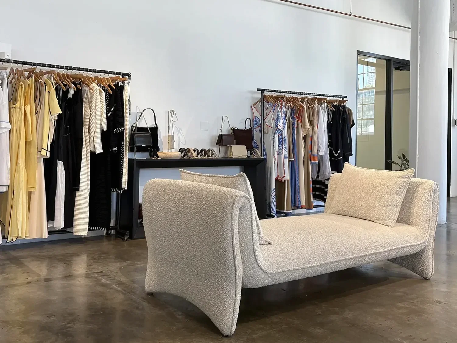 Interior of Place Showroom with racks of clothing, handbags on a table, and a cream-colored textured sofa and chair in the foreground.