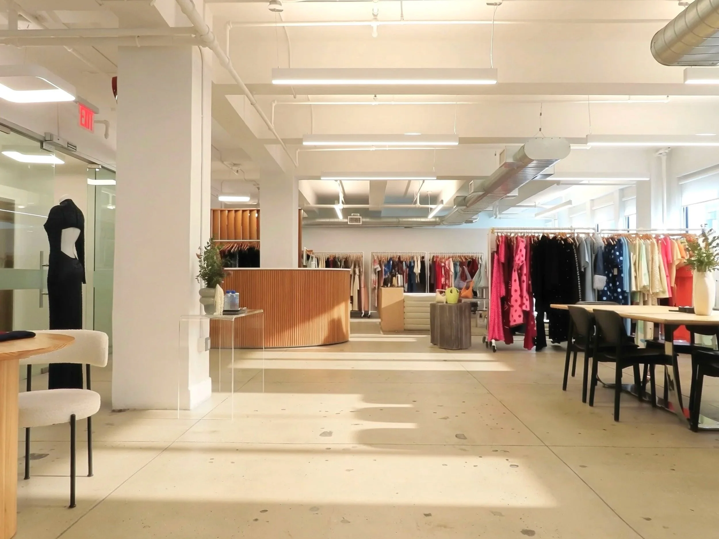 Interior of a clothing store with racks of dresses and a mannequin on the left, illuminated by bright ceiling lights, with a seating area in the foreground.